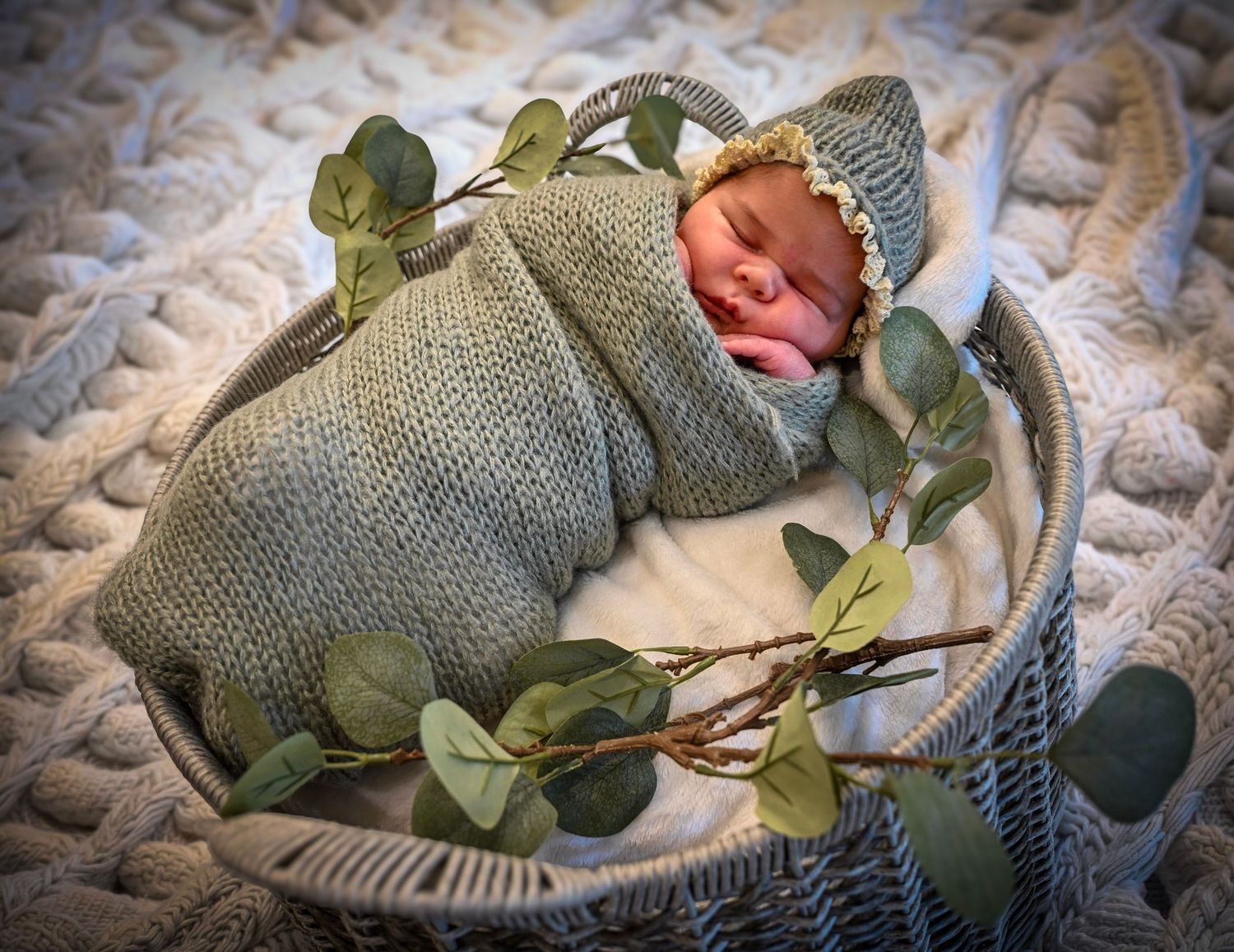 Newborn baby swaddled in sage green knit wrap with bonnet in a white basket decorated with eucalyptus leaves.
