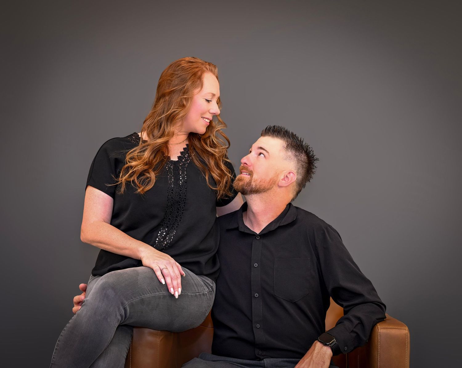 Candid portrait of two people in black attire sharing a moment while seated on brown leather chair.