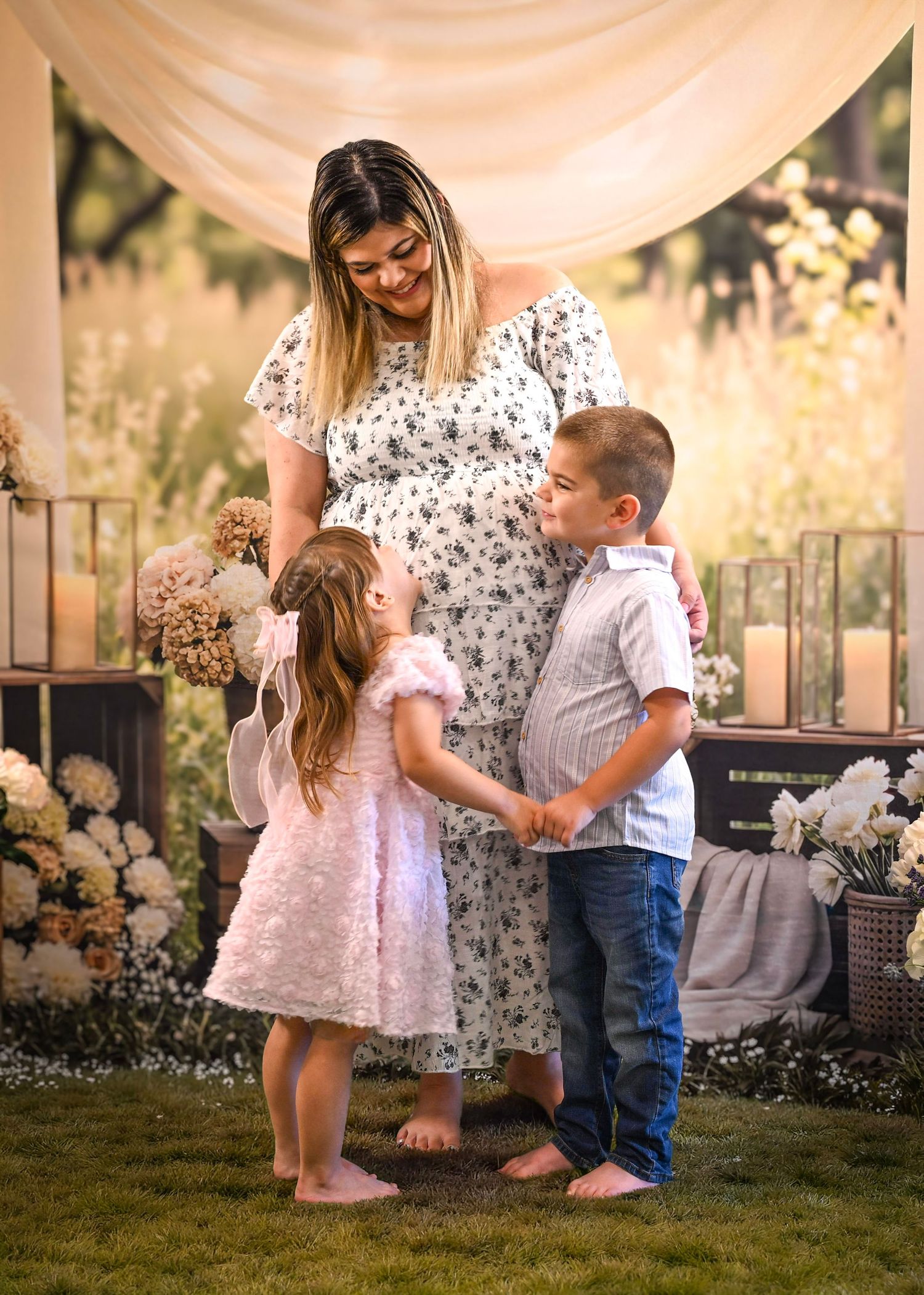 Pregnant woman in floral dress smiling at two young children holding hands during an outdoor maternity photo session.
