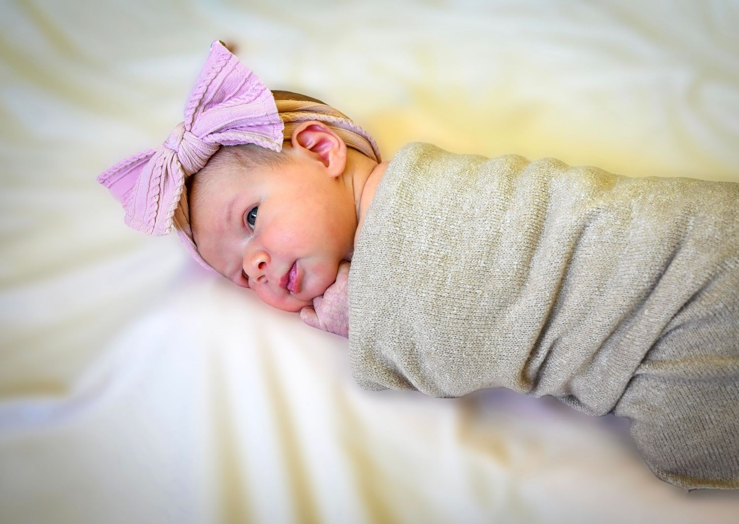 Newborn baby girl swaddled in gray wrap wearing a pink bow headband on white bedding.