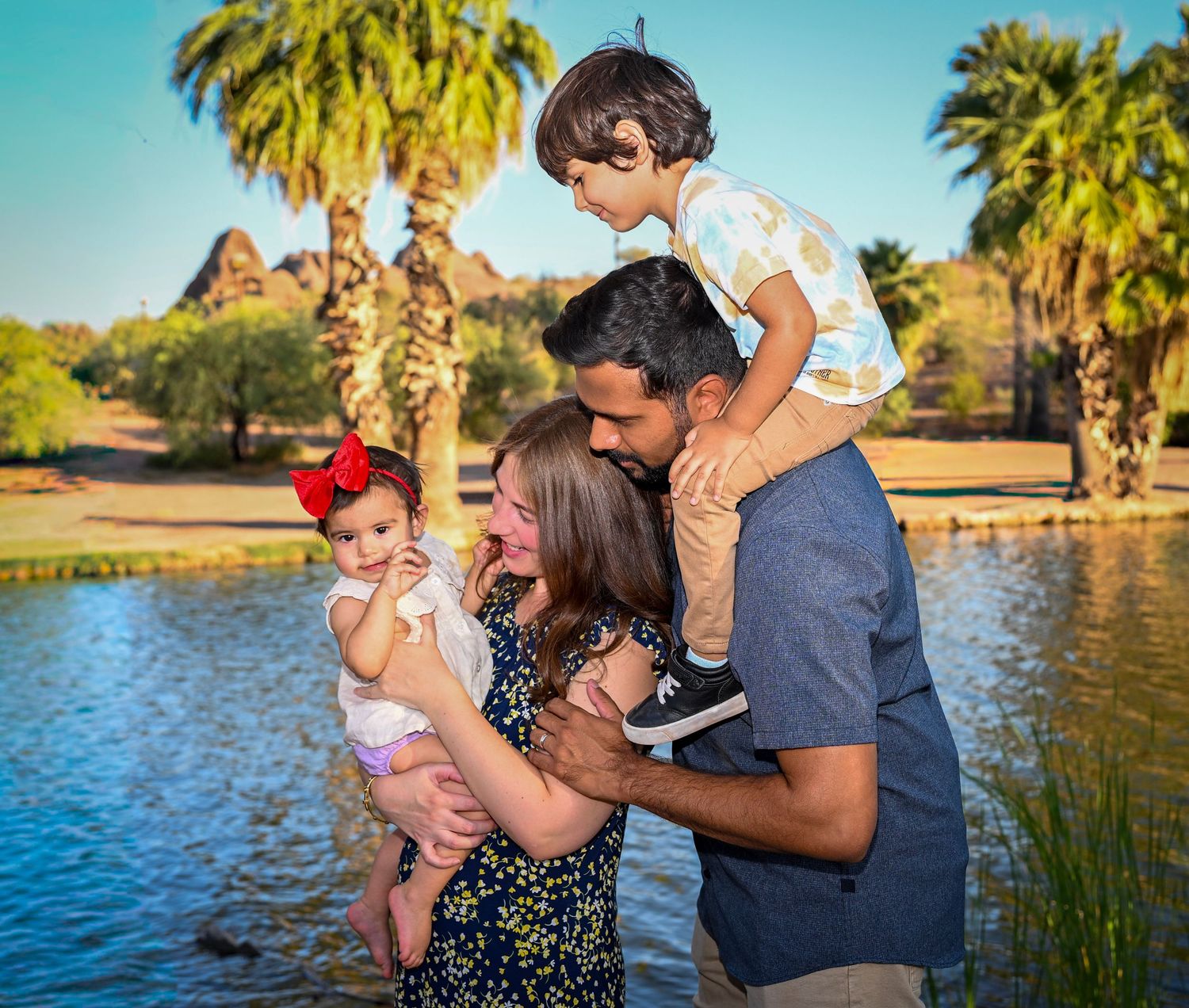 Family of four enjoying a sunny day at a park with palm trees and a pond, parents holding two young children.