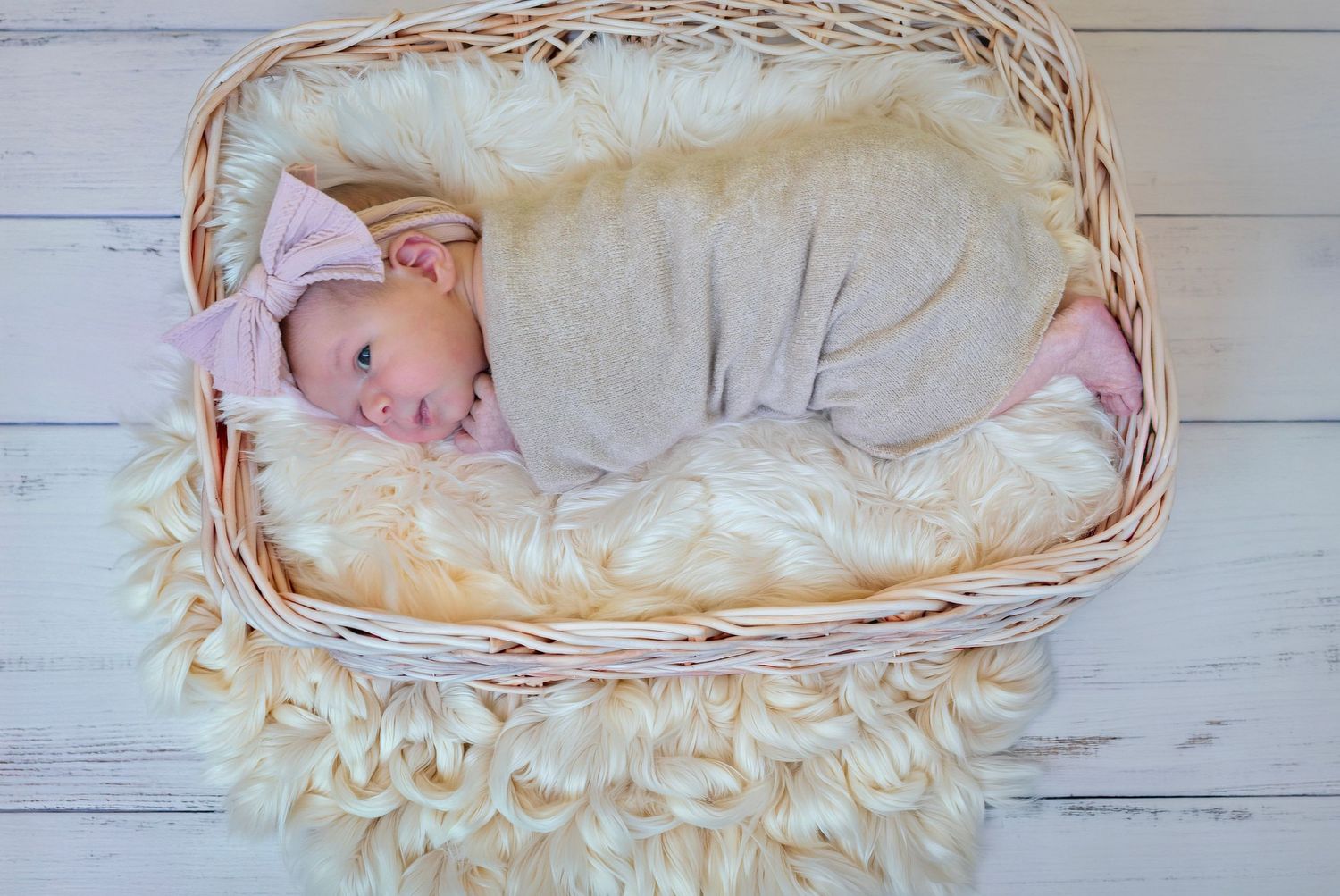 Newborn baby swaddled in gray wrap with pink bow headband sleeping peacefully in a white wicker basket.