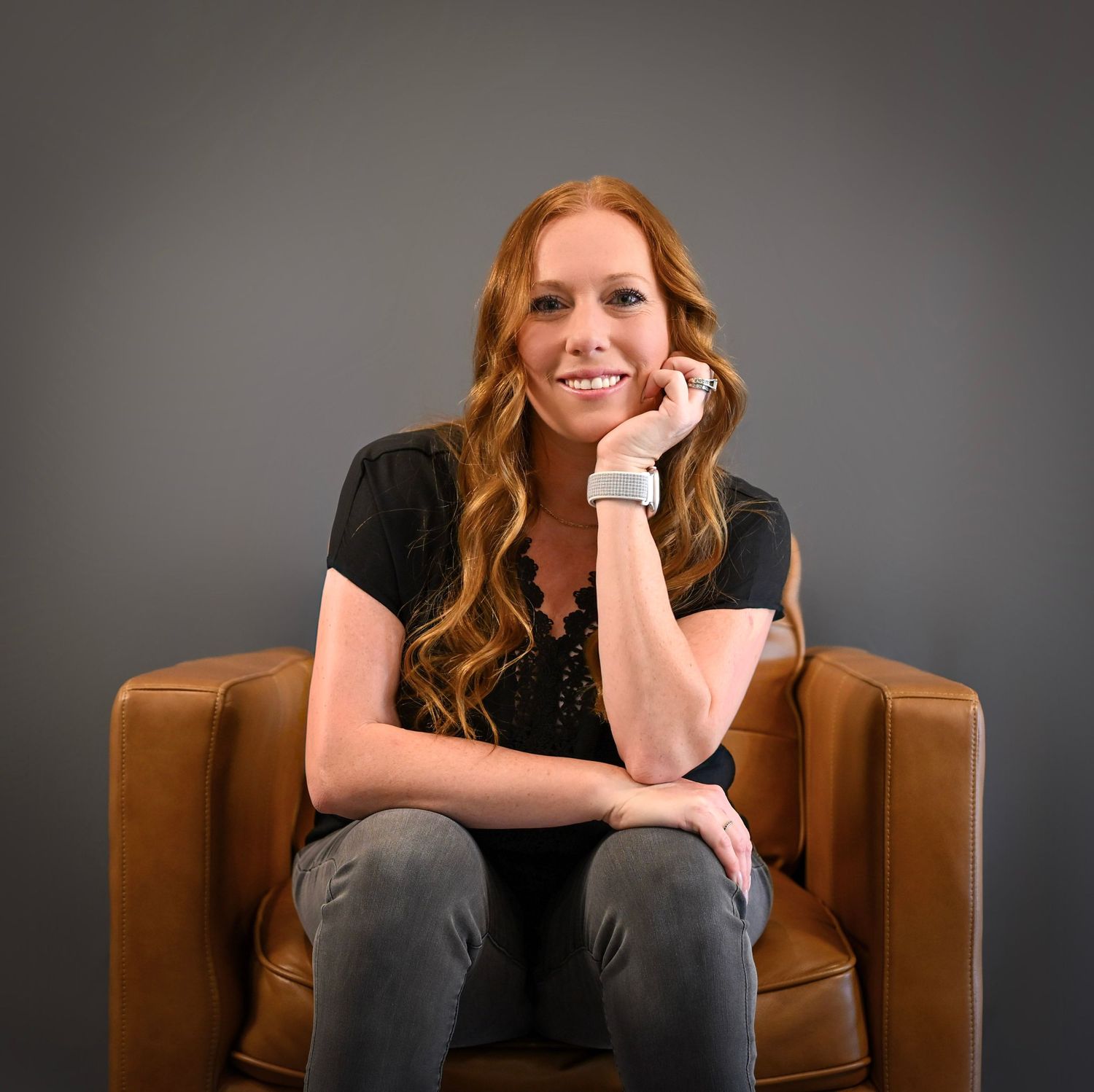 A woman in a black shirt sits casually in a brown leather armchair against a gray background.