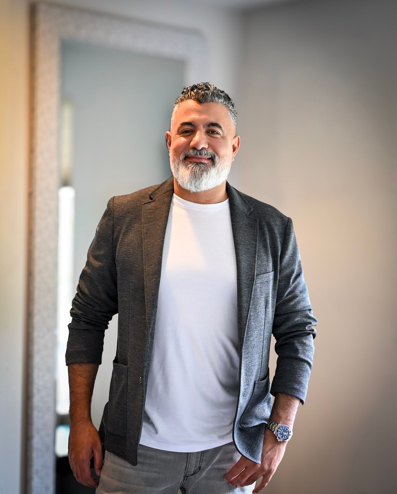 A man in a grey blazer and white t-shirt poses against a light colored wall in a casual business style.