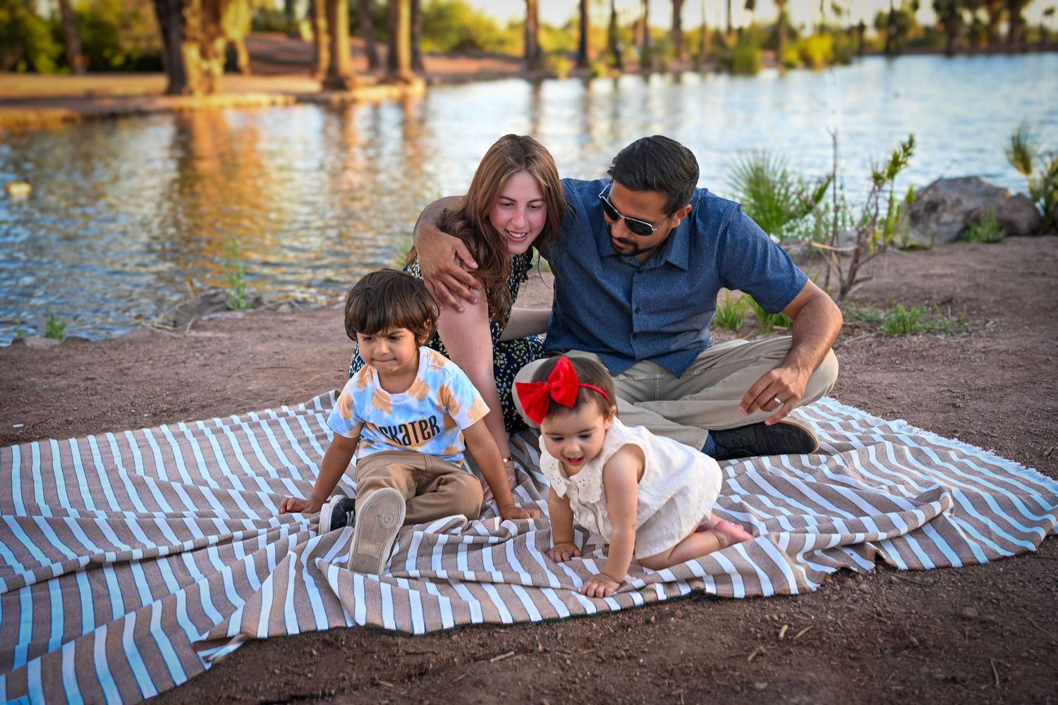 Family of four enjoying a picnic by the lake, with two toddlers playing on a striped blanket outdoors.