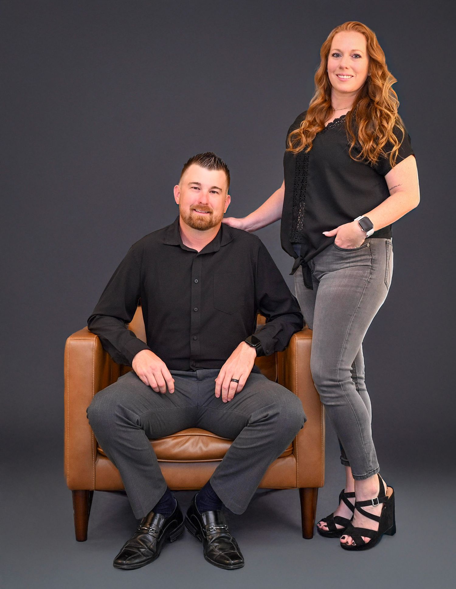 Professional studio portrait of two people in black outfits posing with a brown leather chair against dark background.