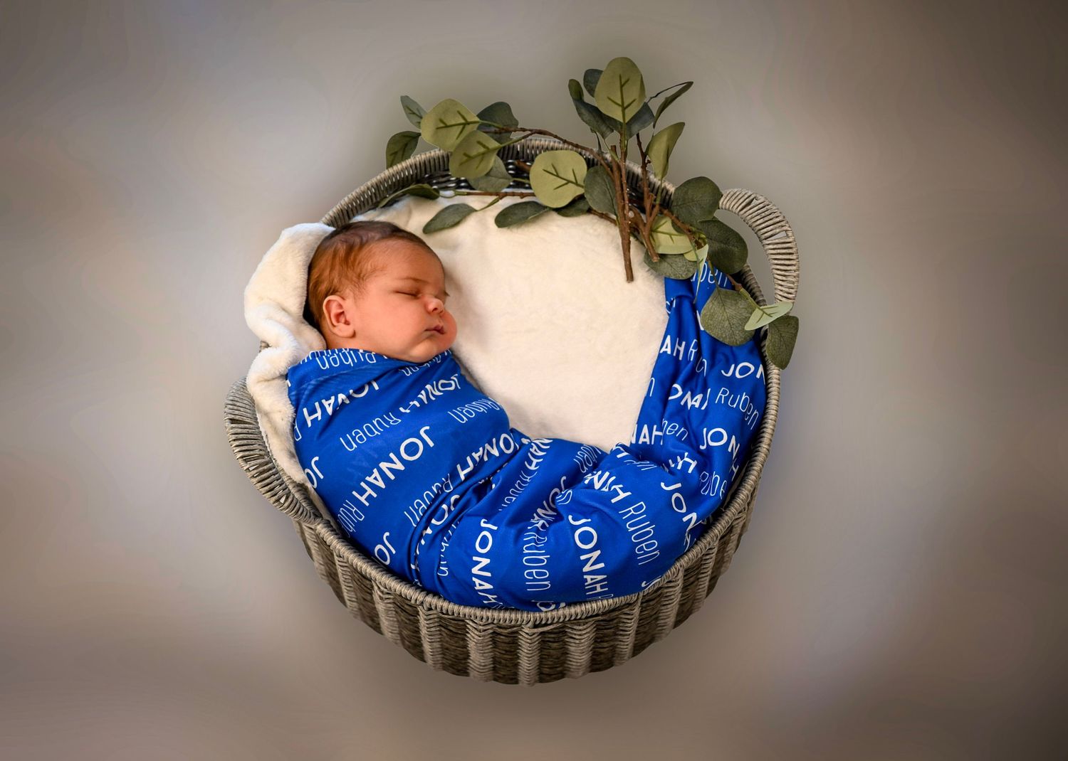 Newborn baby sleeping in a wicker basket wrapped in a blue personalized swaddle blanket with white lettering.