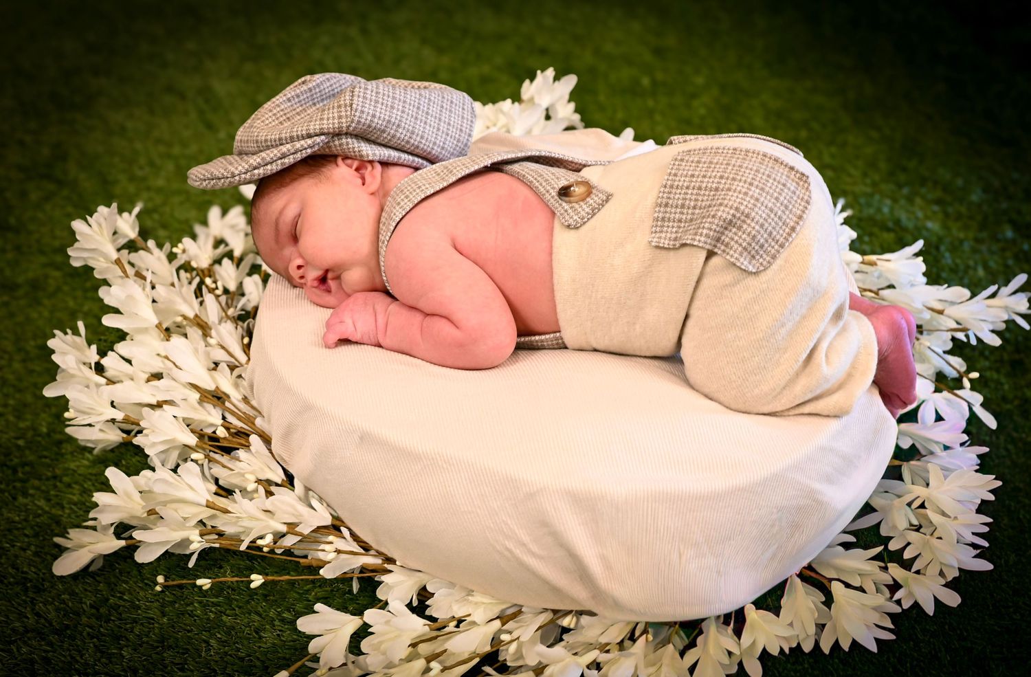 Newborn baby sleeping peacefully in a white bowl surrounded by white flowers, wearing a plaid outfit and bunny hat.