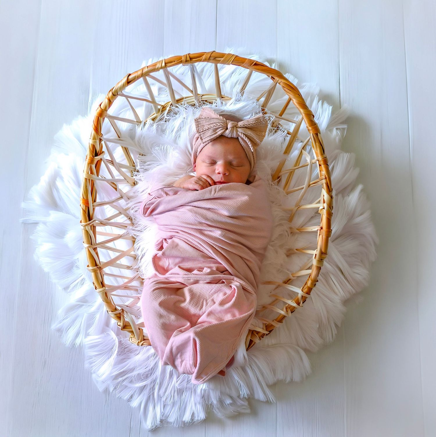 Newborn baby swaddled in pink wrap with bow headband, sleeping in a wicker basket with white fringe liner.