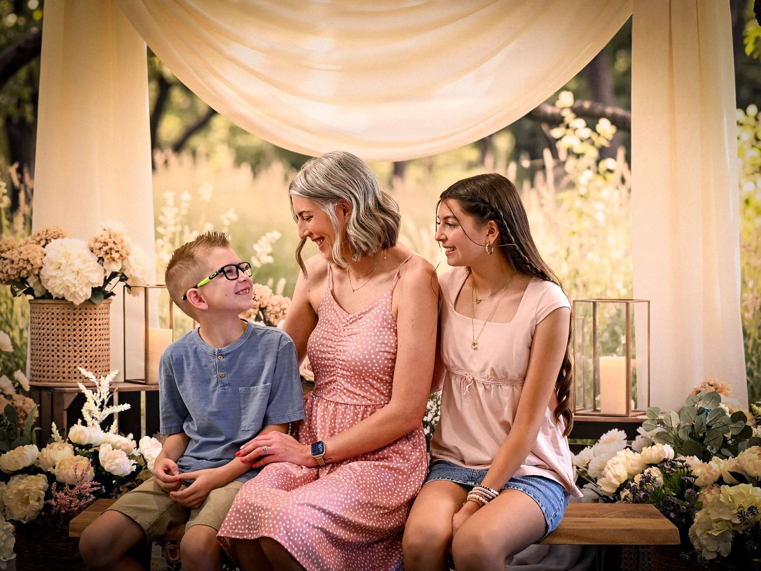 A woman in a pink dress sits on a bench with two children, smiling together amid floral decor in a warm outdoor setting.