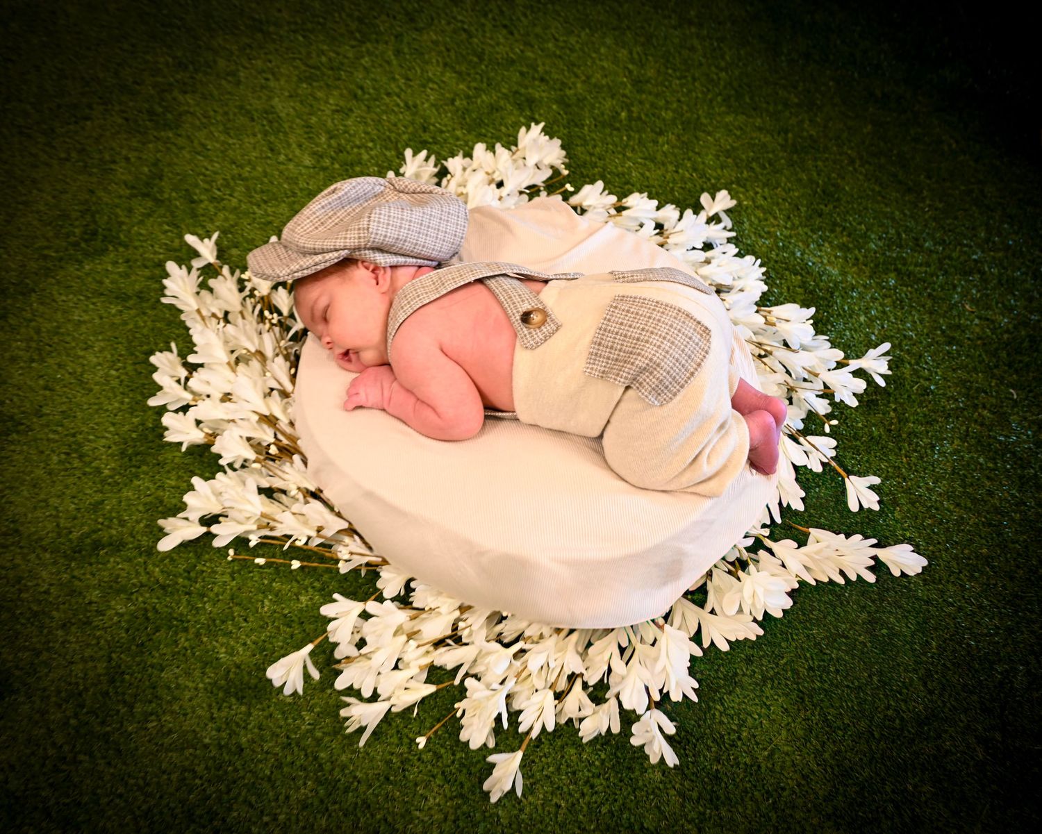 Newborn baby sleeping on white cushion surrounded by white flowers wearing elephant costume on green grass.