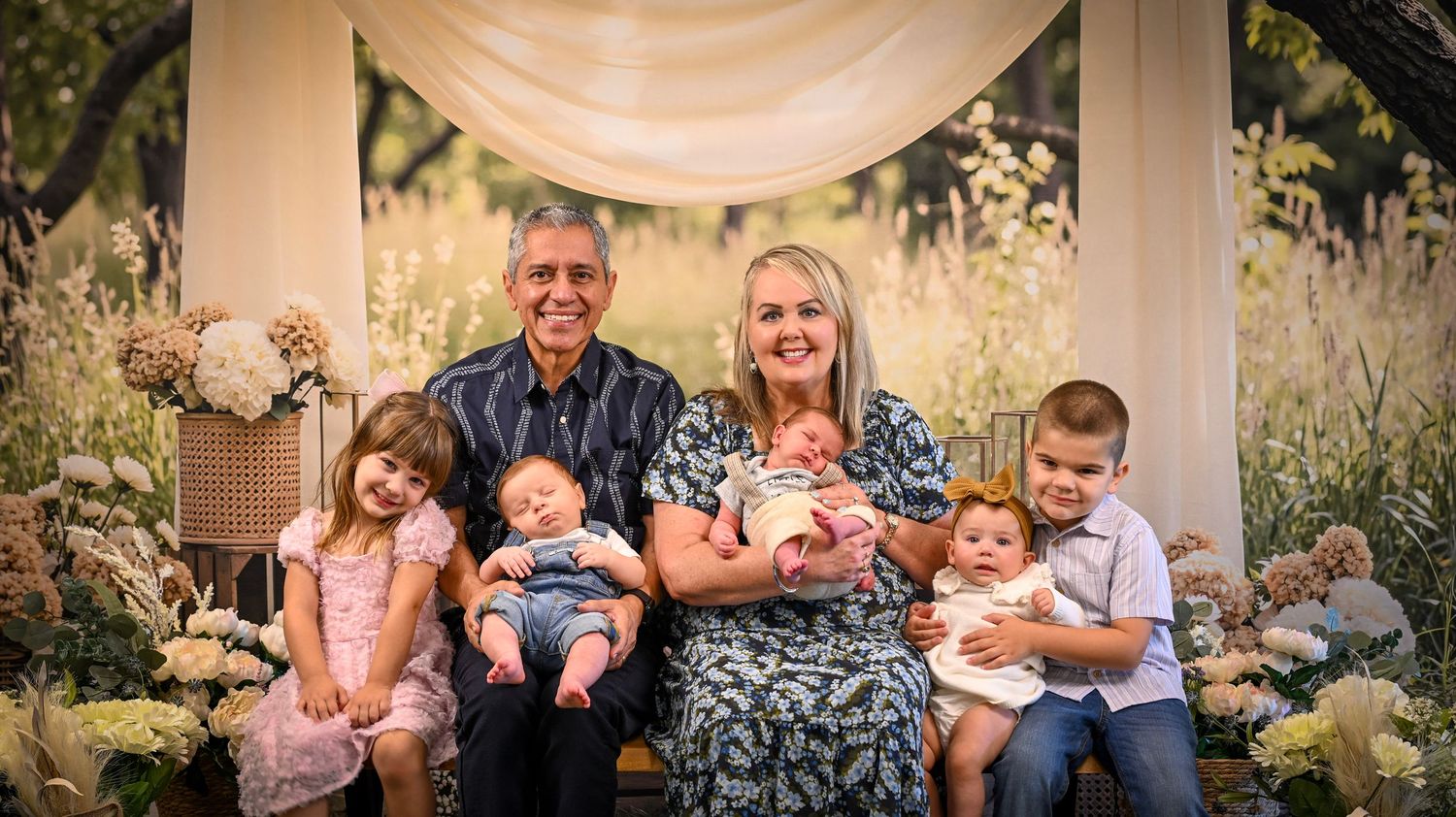 Grandparents pose outdoors with four young grandchildren, holding babies and toddlers in a beautiful floral garden setting.