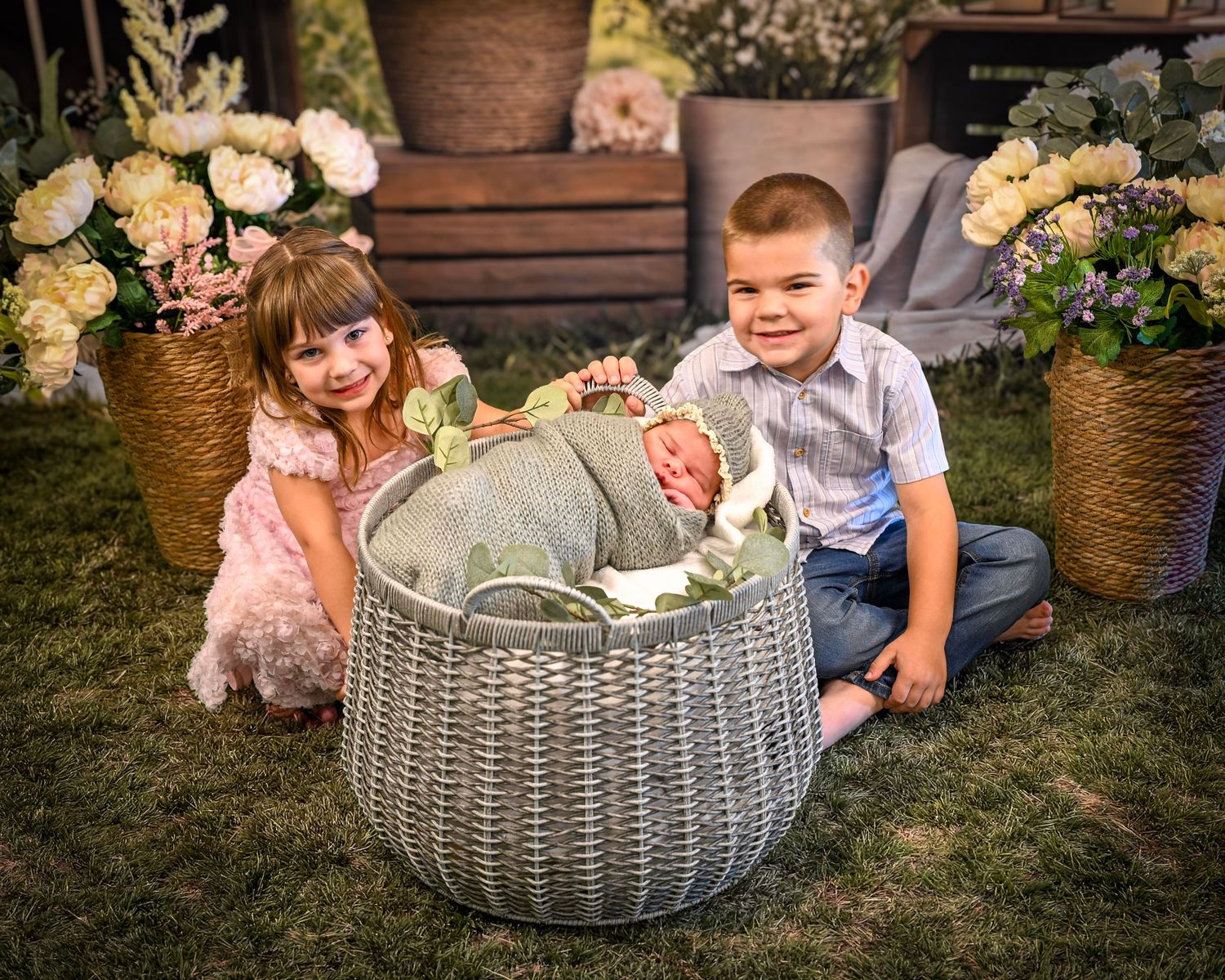 Two children sit on grass beside a wicker basket holding a newborn baby, surrounded by floral arrangements.