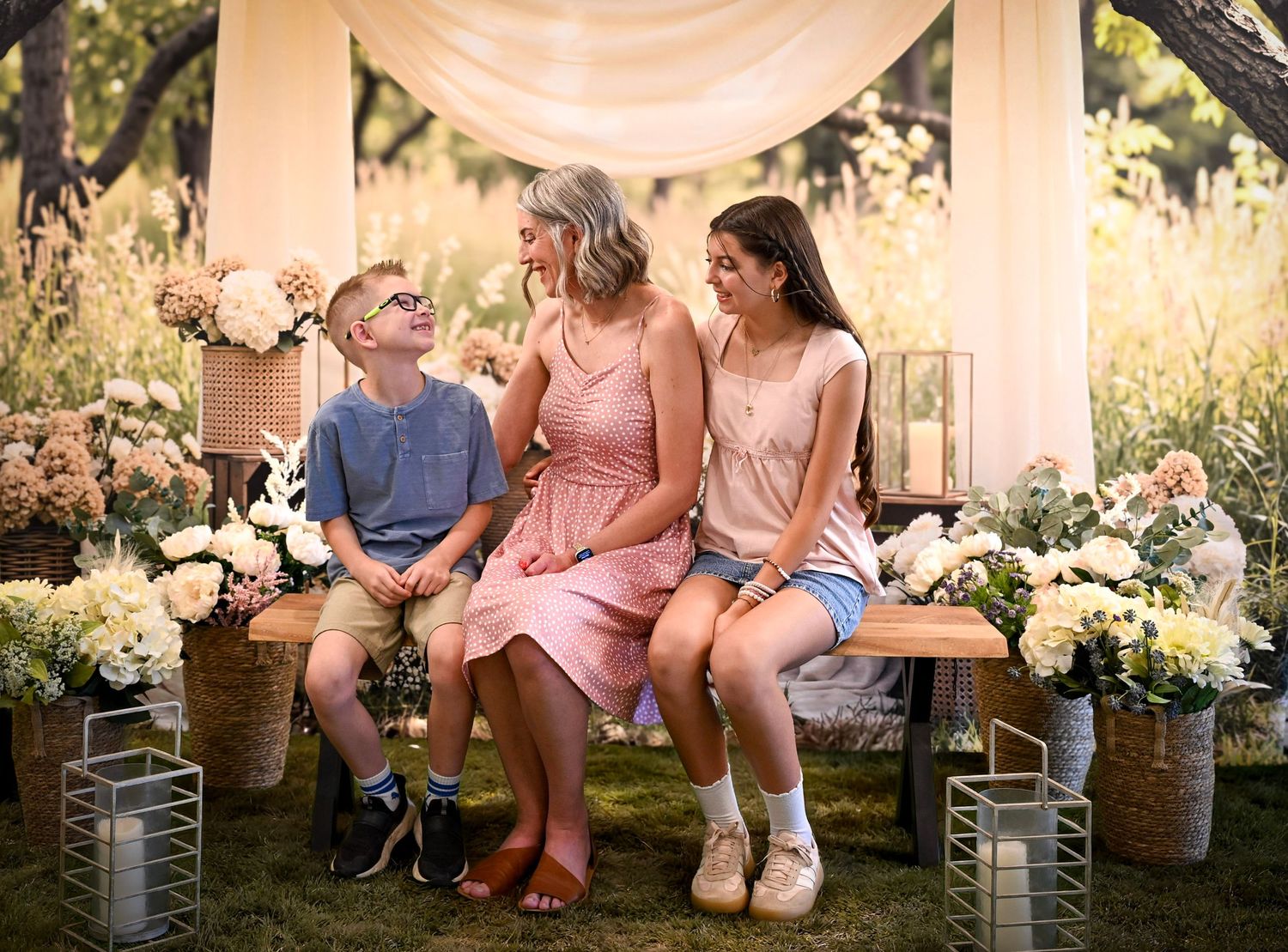 A woman in pink sits on a bench with two children, surrounded by white flowers in a garden setting.