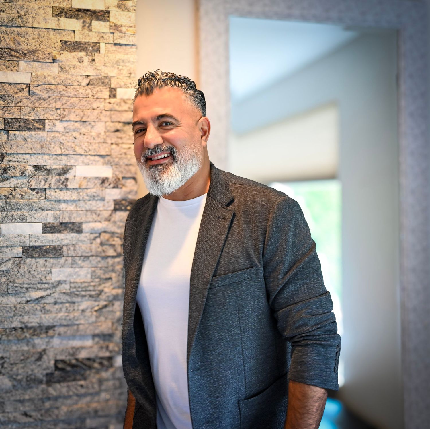 Professional portrait against a textured stone wall background wearing a grey blazer and white t-shirt.