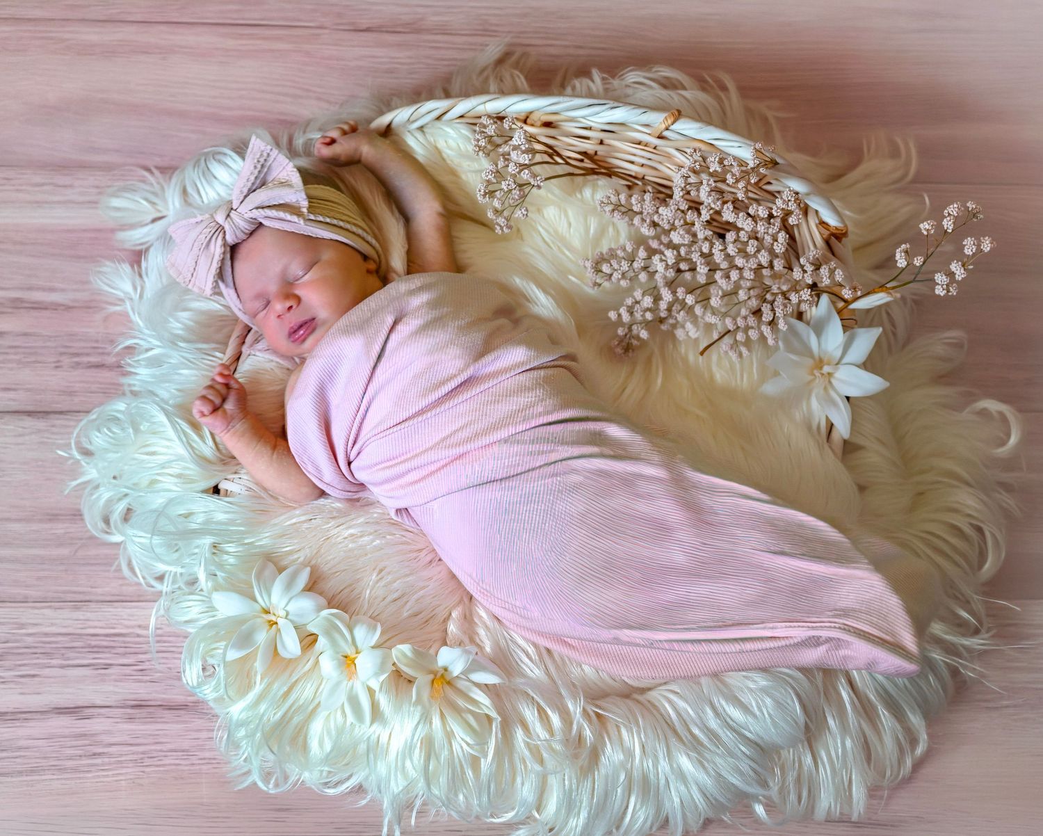 Newborn baby sleeping peacefully wrapped in pink swaddle with bow headband on fluffy white fur nest with flowers.