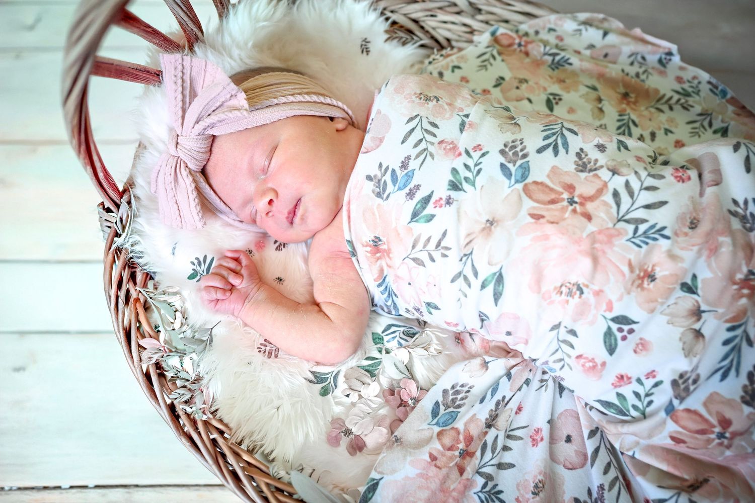 Sleeping newborn baby wrapped in floral blanket with pink bow headband resting in wicker basket.