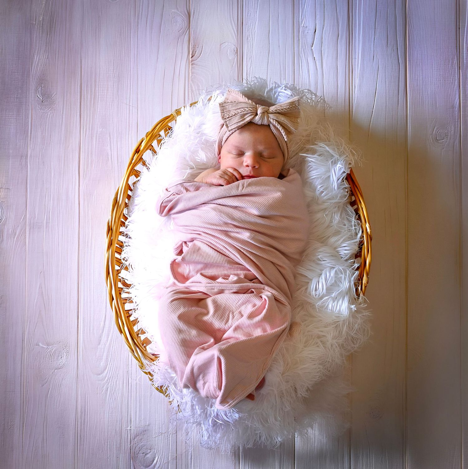 Sleeping newborn baby girl swaddled in pink wrap with bow headband resting in wicker basket with white fur.