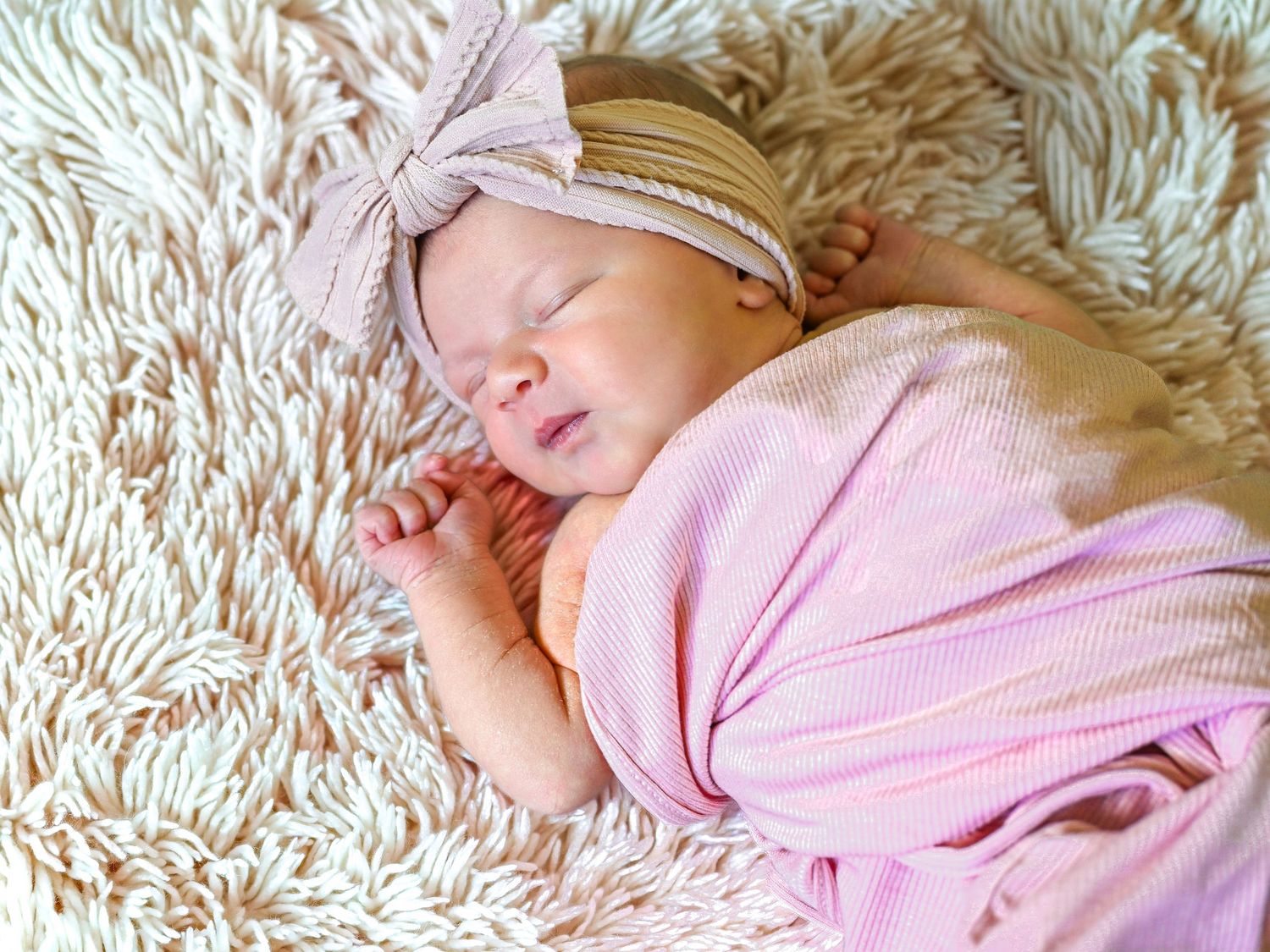 Peaceful newborn baby sleeping swaddled in pink striped blanket with bow headband on fluffy white rug.
