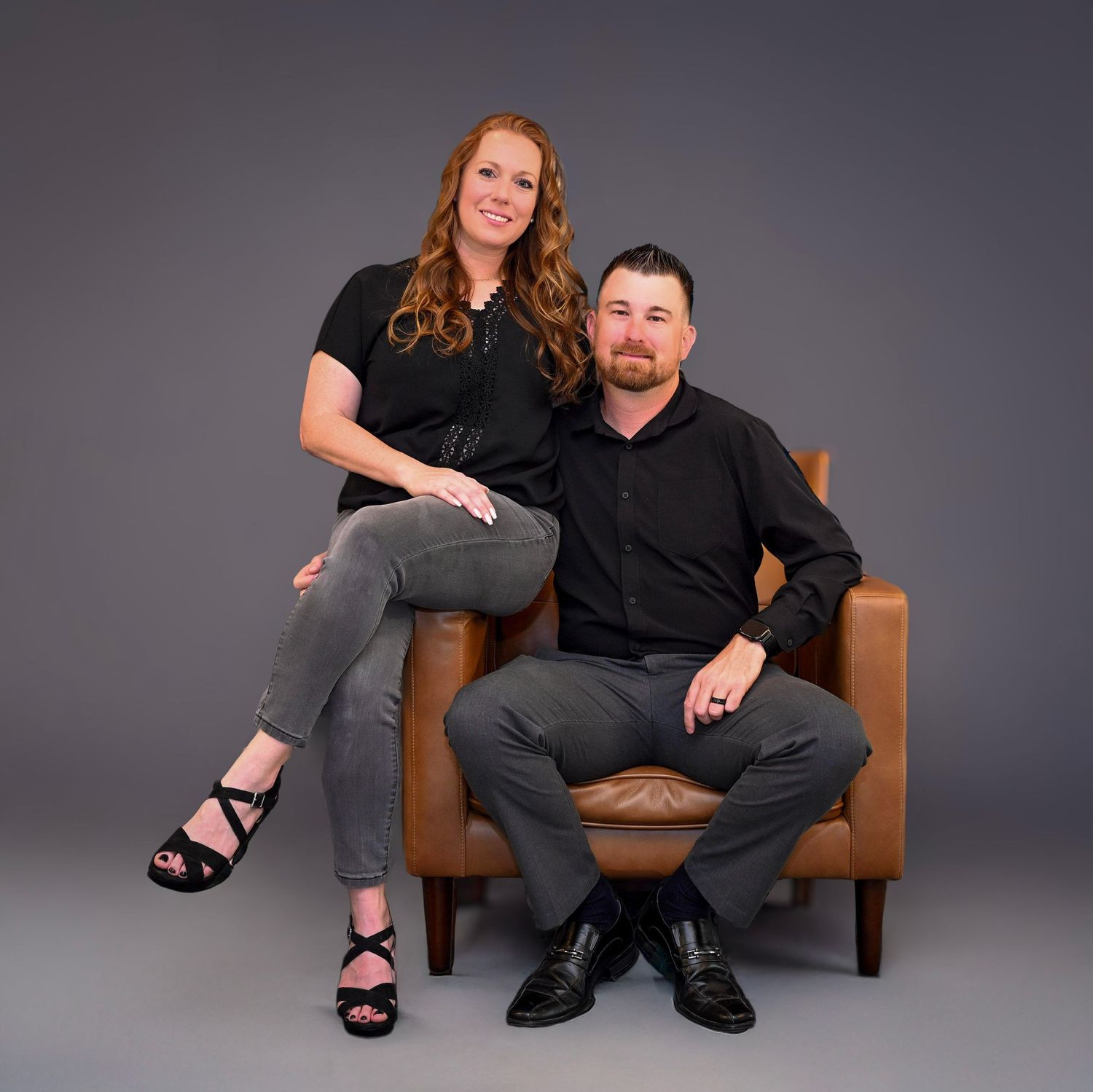A couple poses together on a brown leather chair during a professional studio photoshoot against a gray background.
