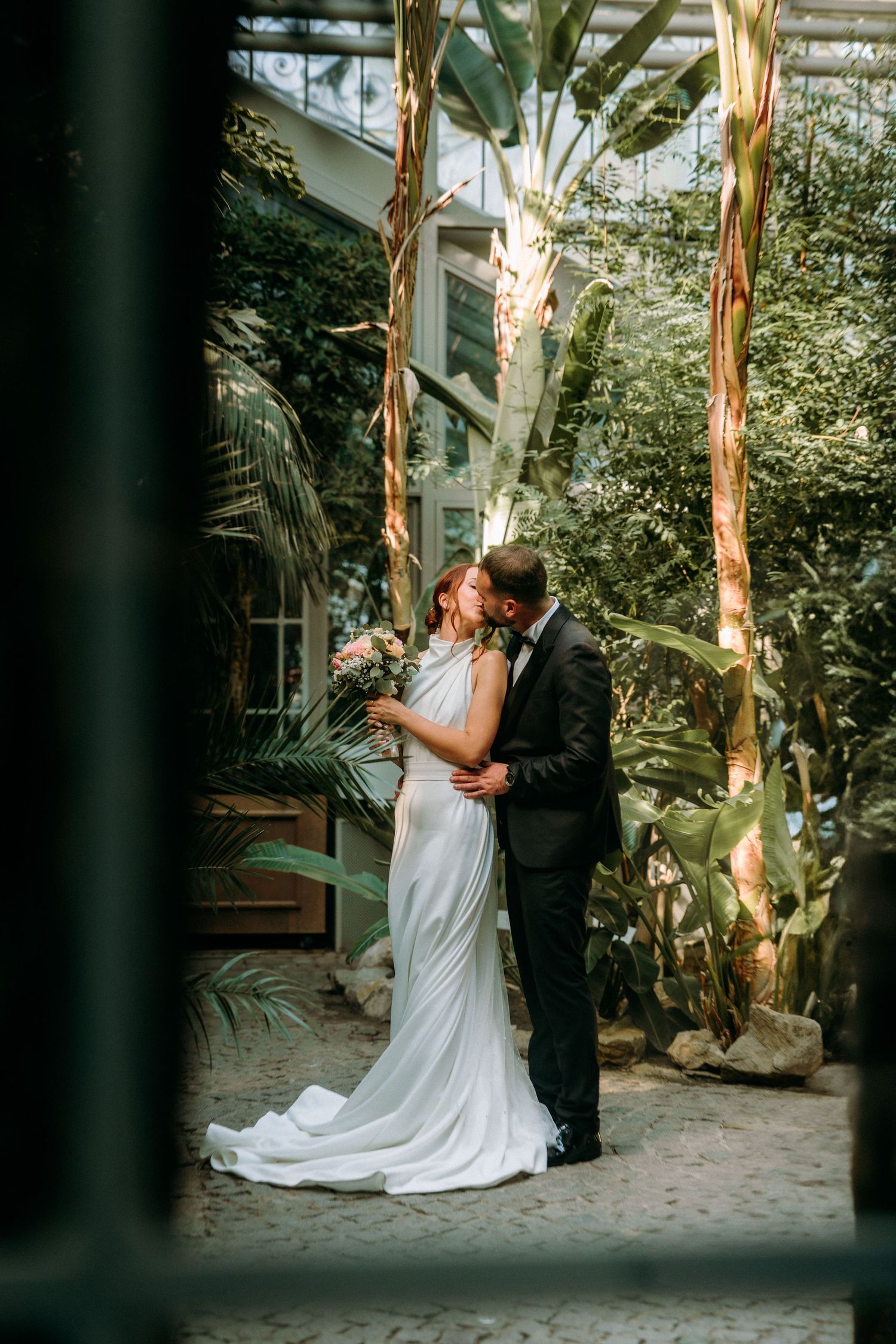 Romantic wedding couple embracing in a tropical greenhouse setting with natural light filtering through.