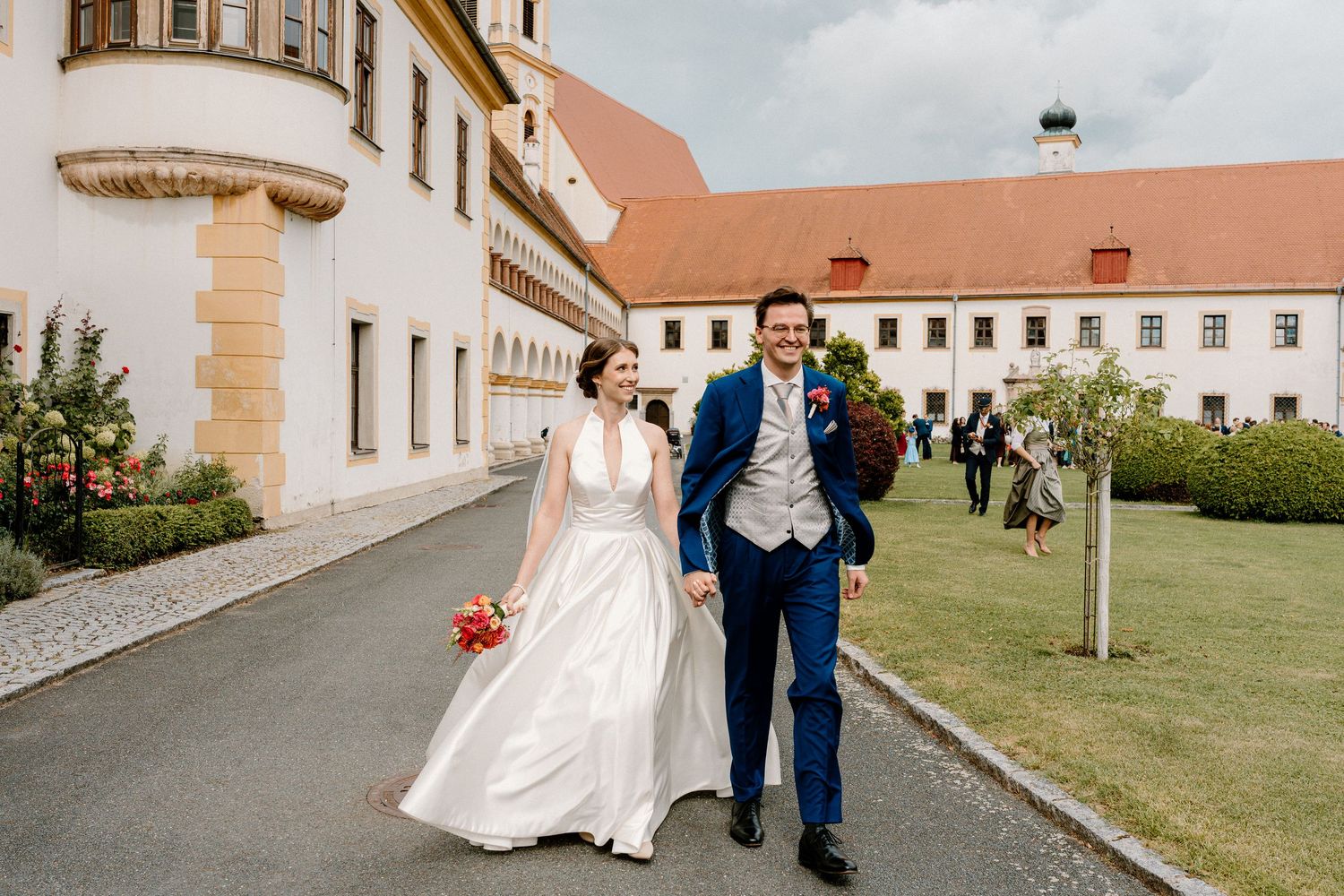 Wedding couple walks along a path beside a white building with red roof tiles.