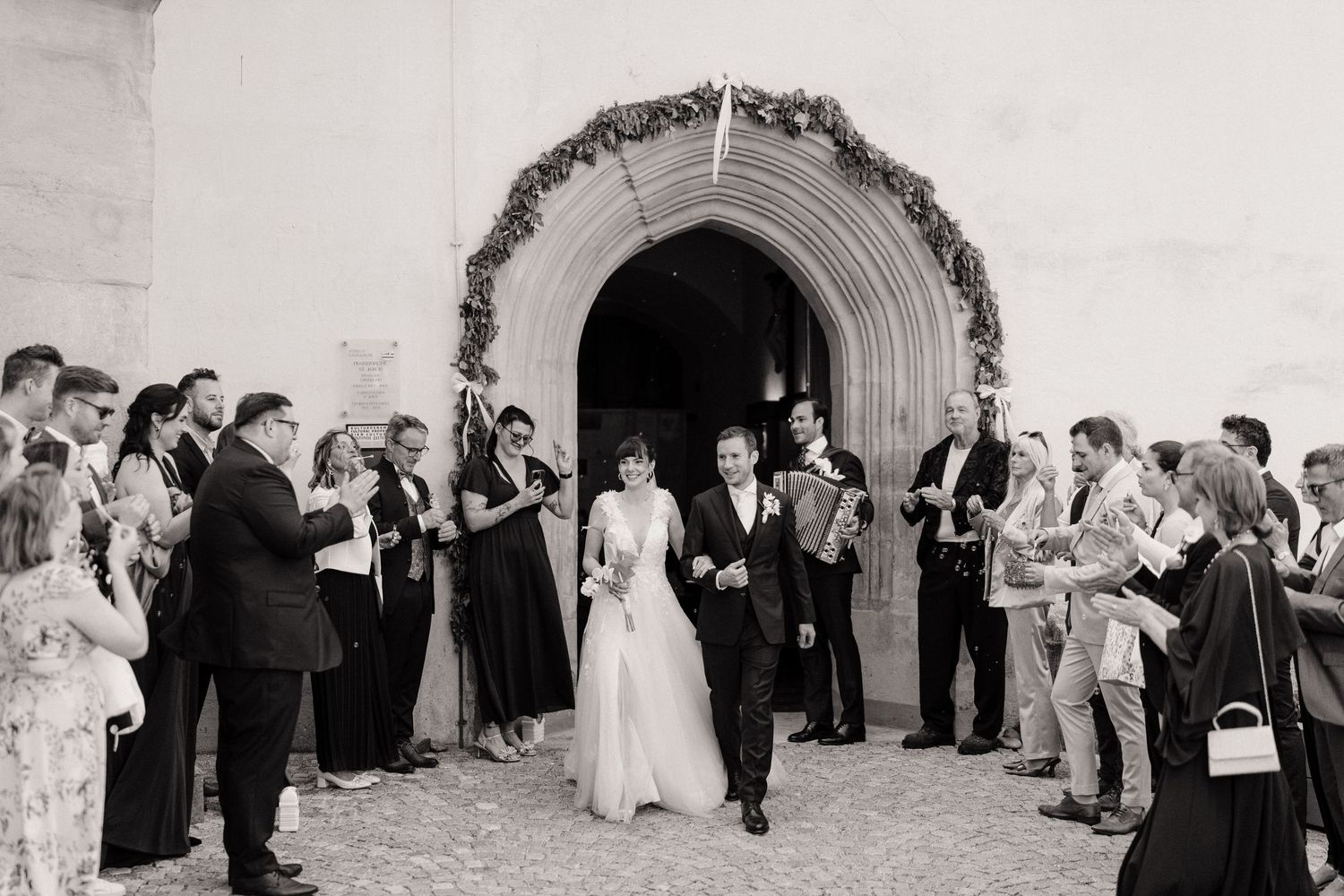 Bride and groom exit church through floral arch, greeted by guests in black and white wedding photo.