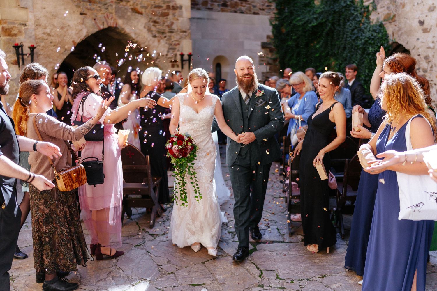Bride and groom walk down aisle as guests toss colorful confetti at outdoor stone wedding venue.