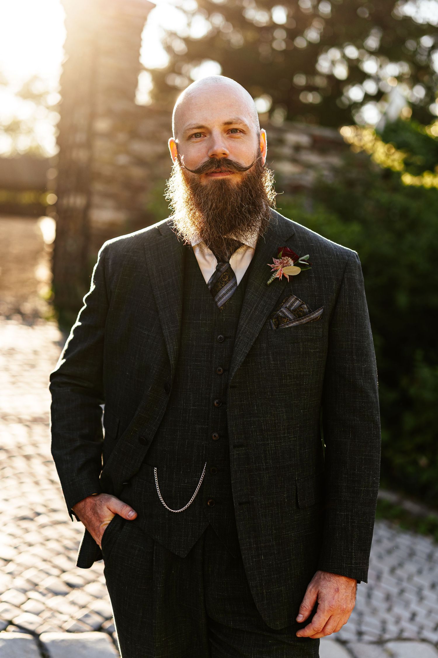 Bald groom with long beard in dark plaid three-piece suit with boutonniere and pocket square poses outdoors.