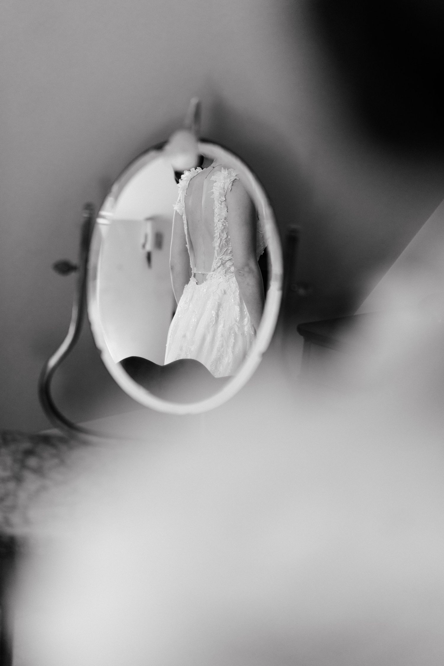 Black and white photo of a wedding dress reflected in an oval mirror, with a blurred figure in the foreground.