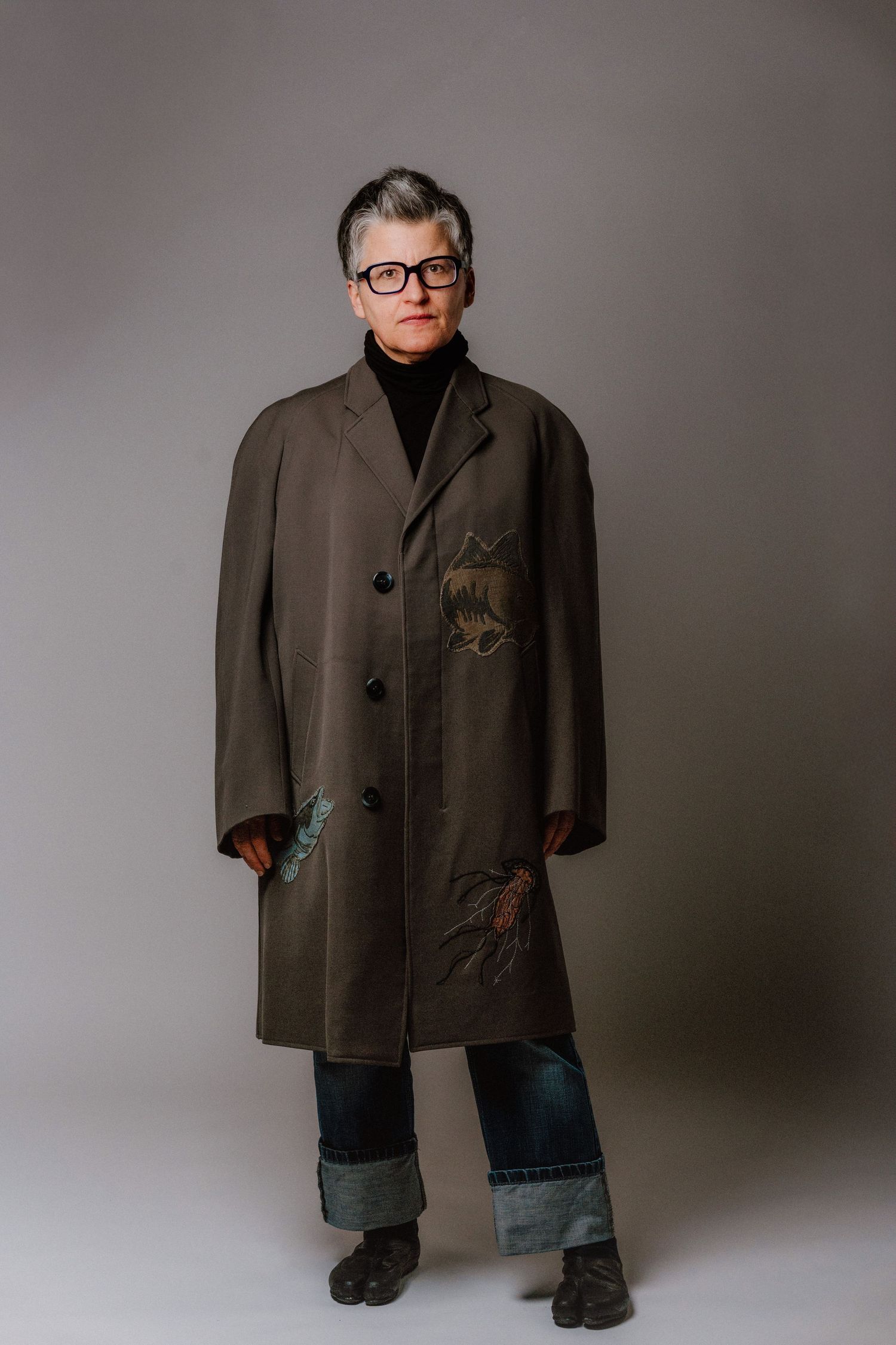 Person wearing olive green embroidered coat, jeans, and glasses standing against gray background.