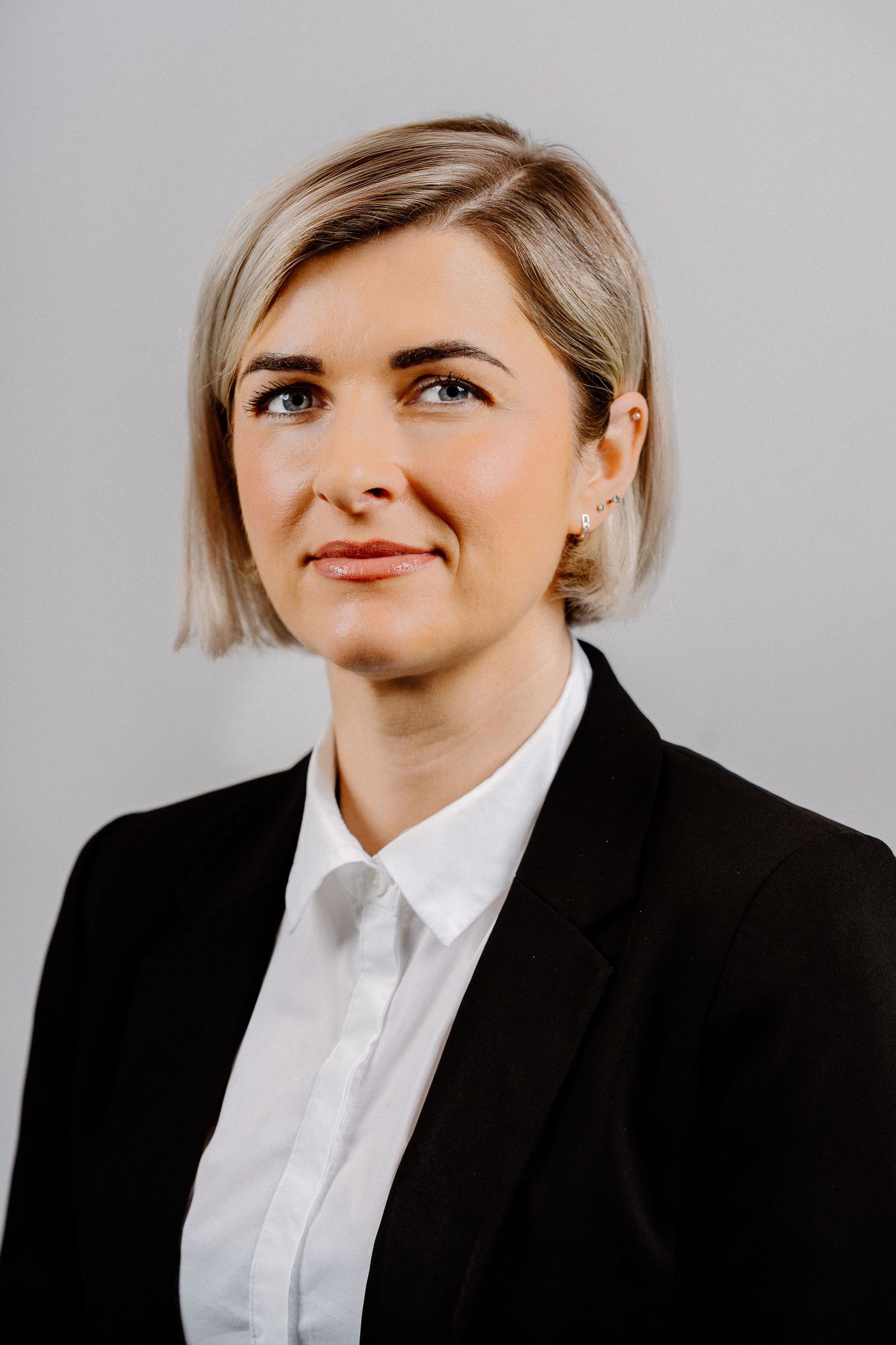 Professional headshot of a blonde woman in a black blazer and white shirt, smiling confidently against a gray background.