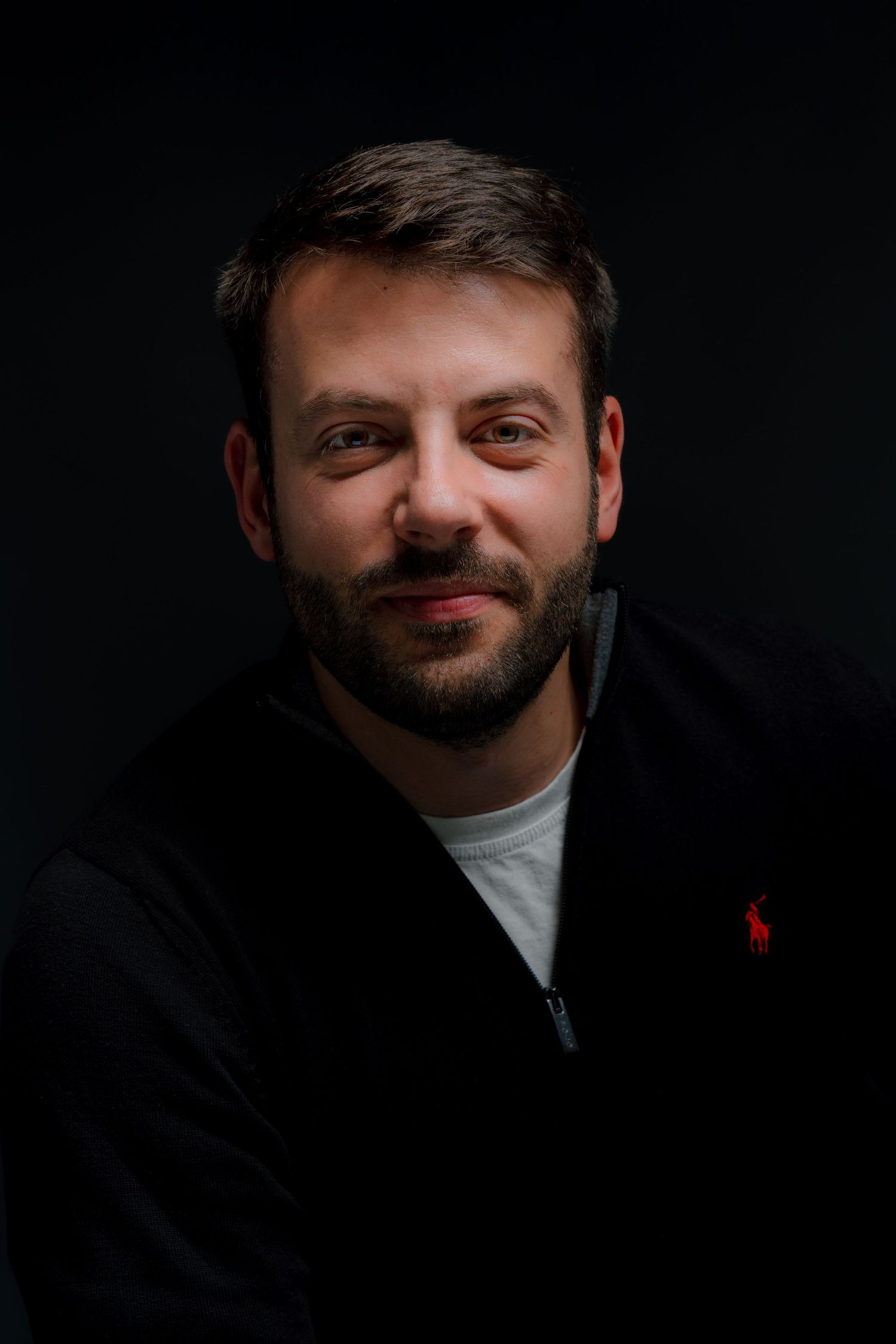 Man with beard smiling, wearing black Ralph Lauren zip-up hoodie, portrait shot against dark background.