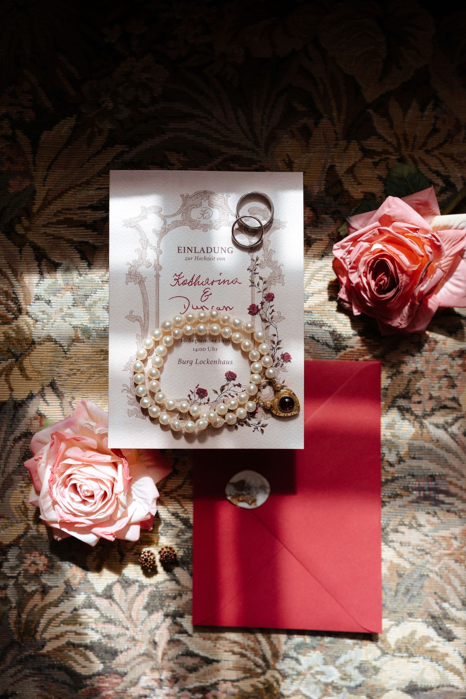 Elegant wedding invitation with pearl bracelet, gold rings, pink roses, and red envelope on floral background.