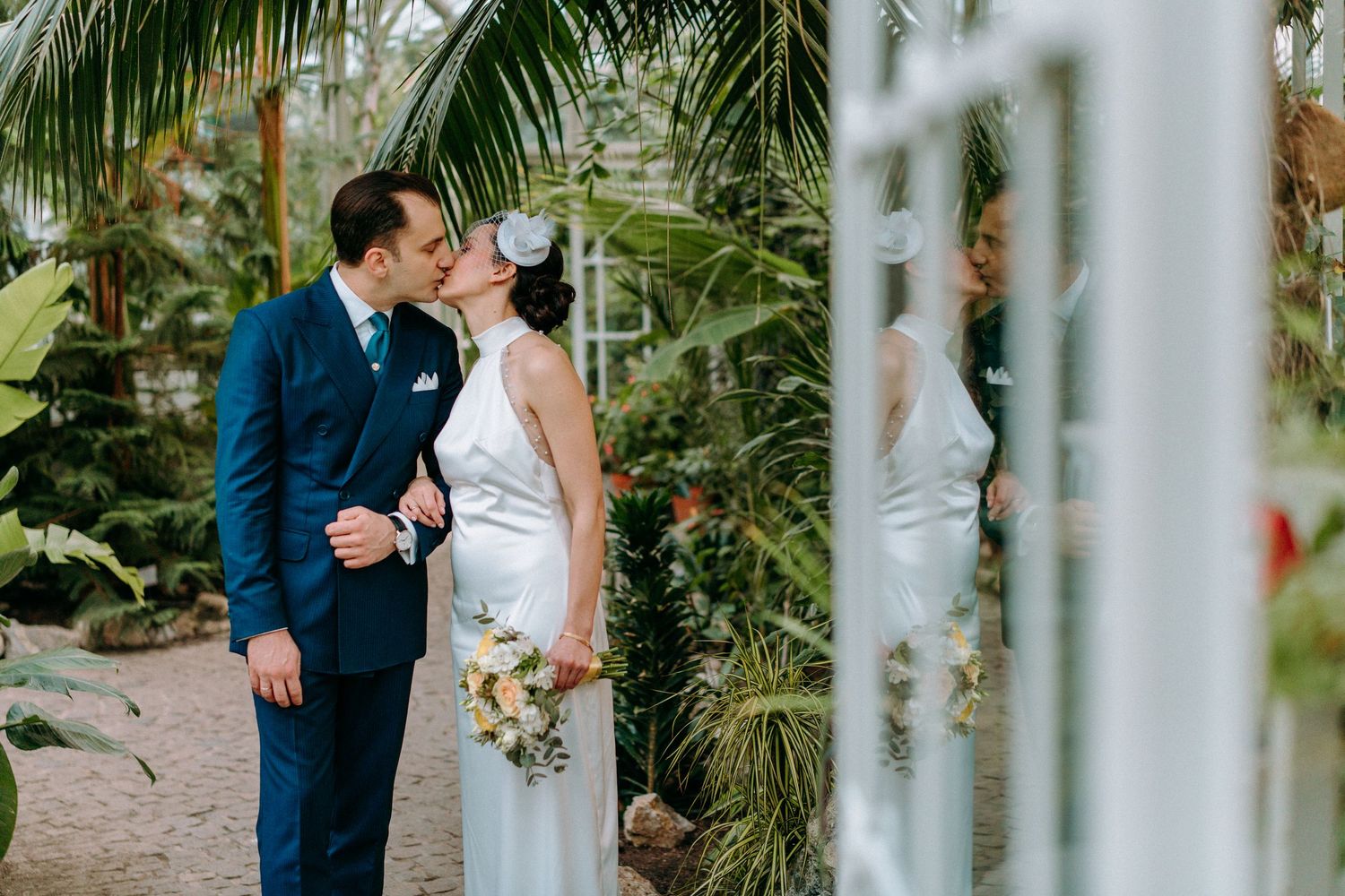 Newlyweds embrace in a garden conservatory with white pillars and tropical foliage.