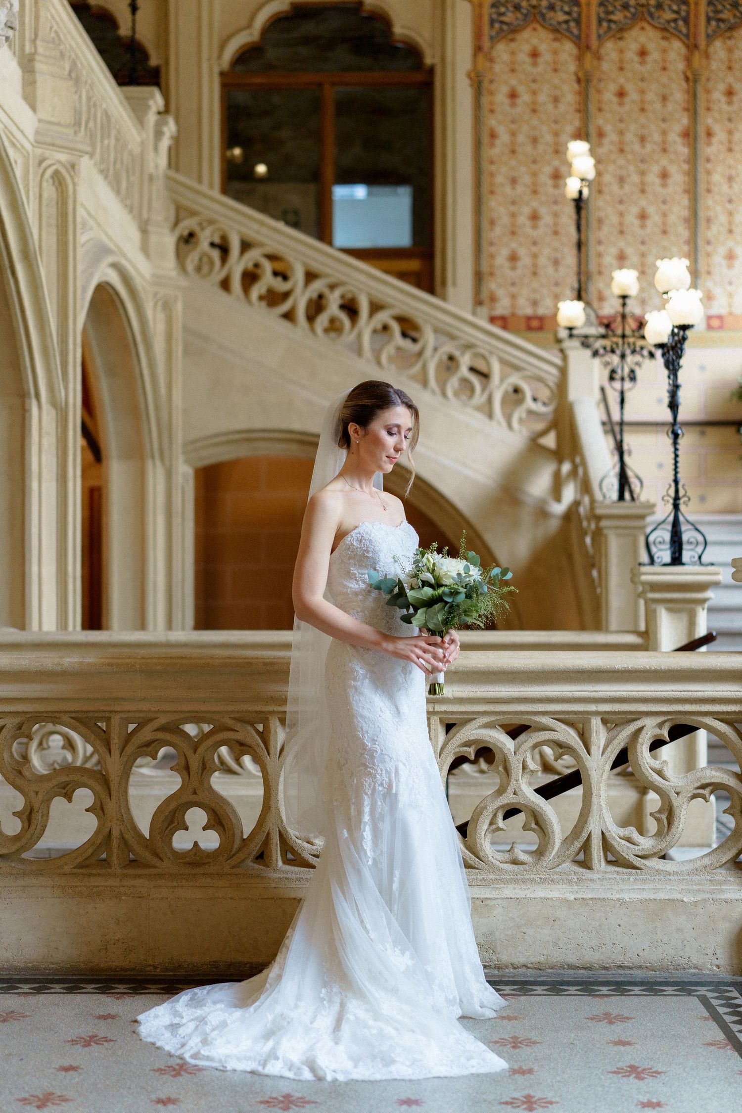A bride in a fitted lace gown poses with her bouquet on ornate stone stairs.