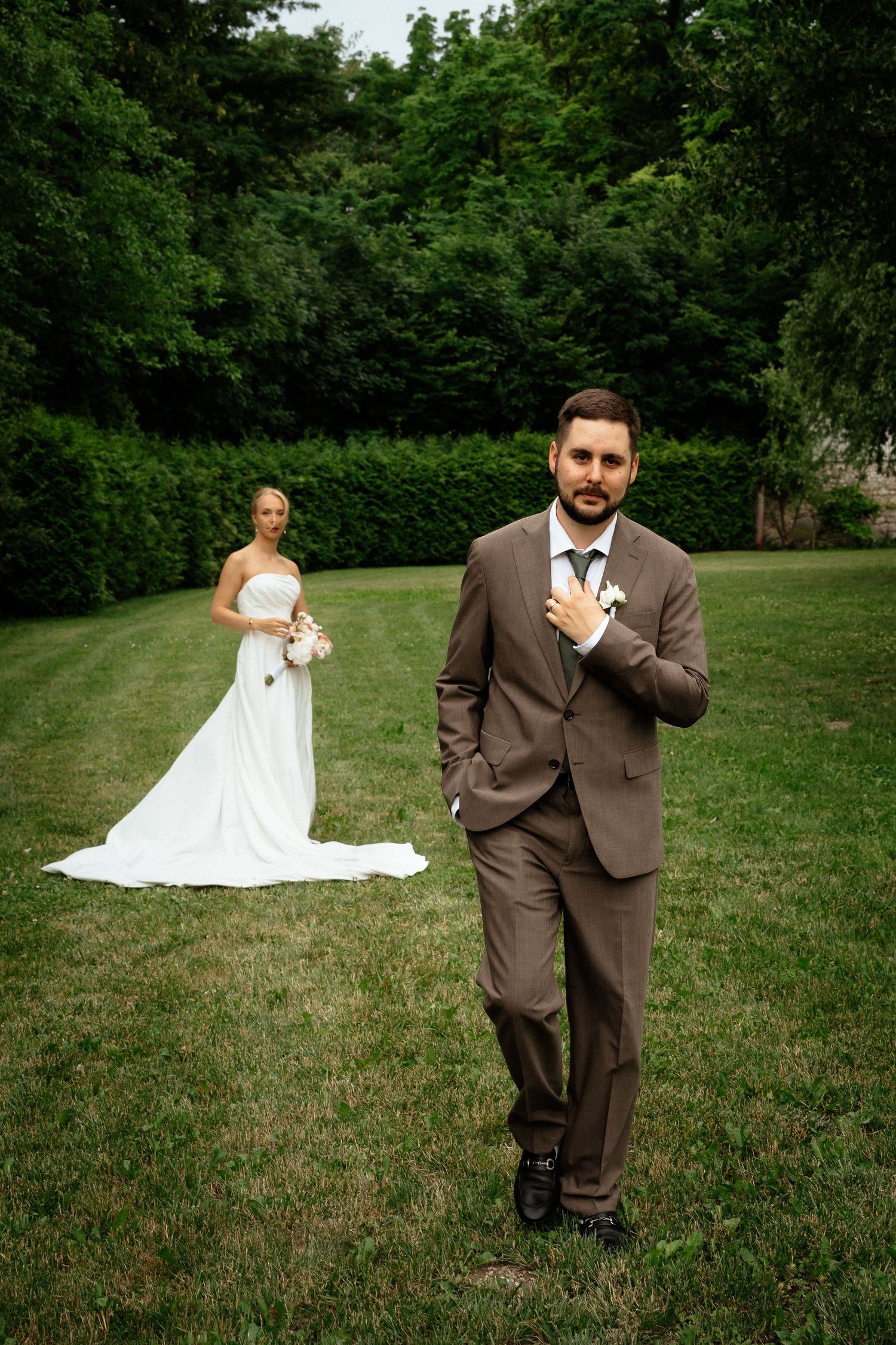 Groom in brown suit stands in foreground while bride in white gown holds bouquet on green lawn surrounded by trees.