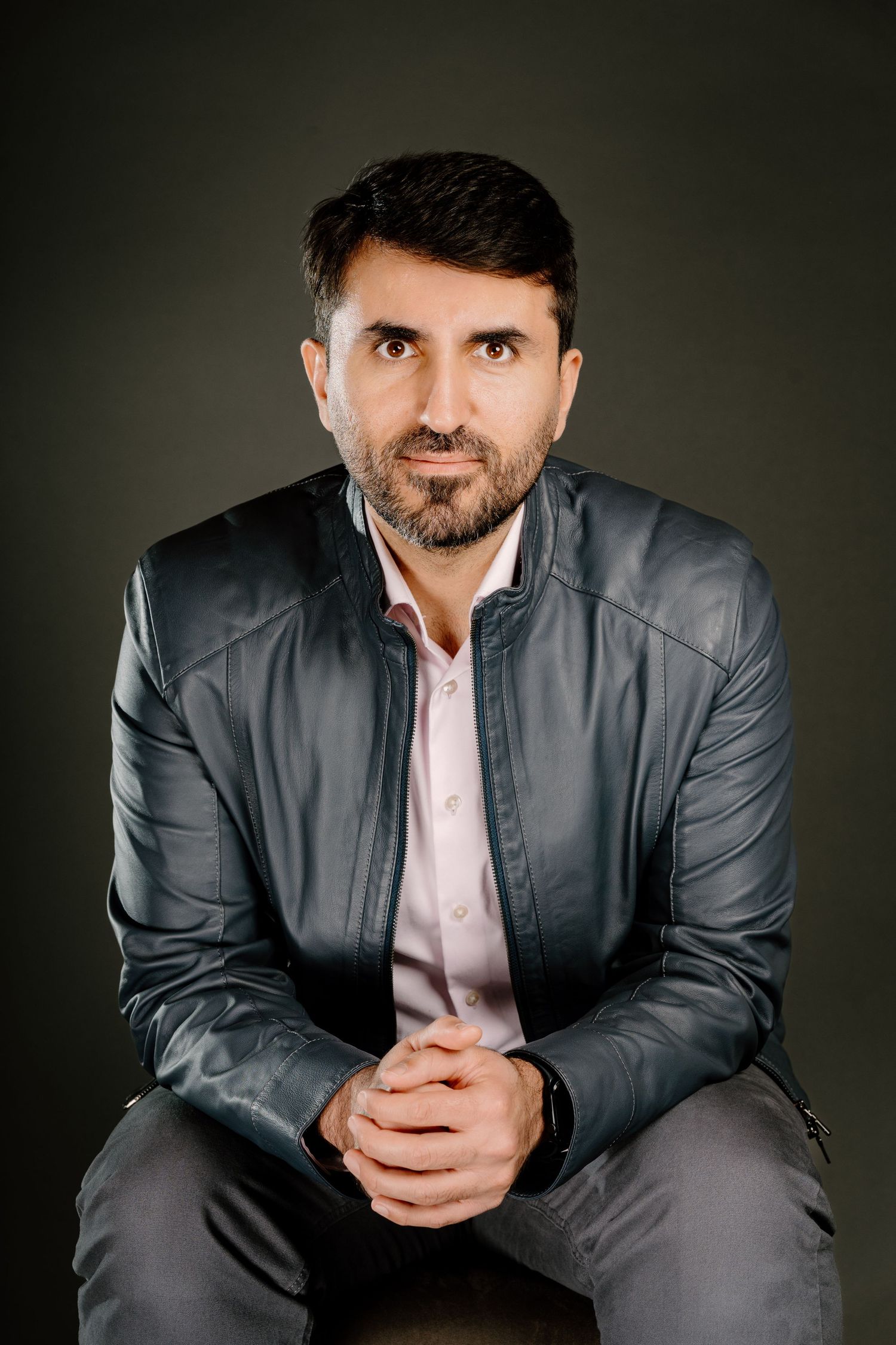 A bearded man in a gray leather jacket sits with clasped hands, posing confidently against a dark background.