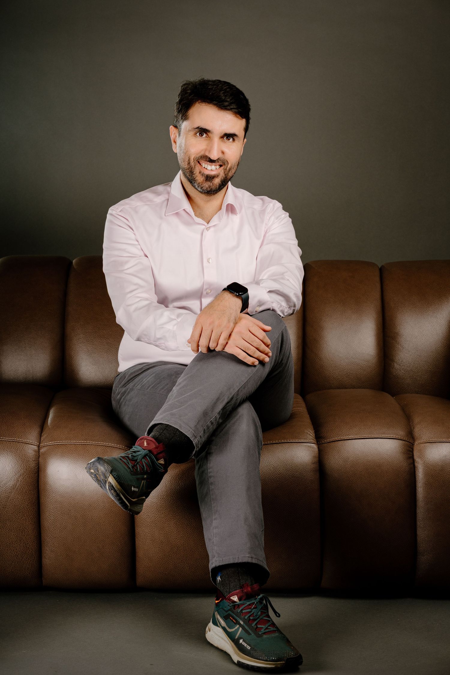 A smiling bearded man in a pink shirt sits relaxed on a brown leather sofa with legs crossed wearing sneakers.