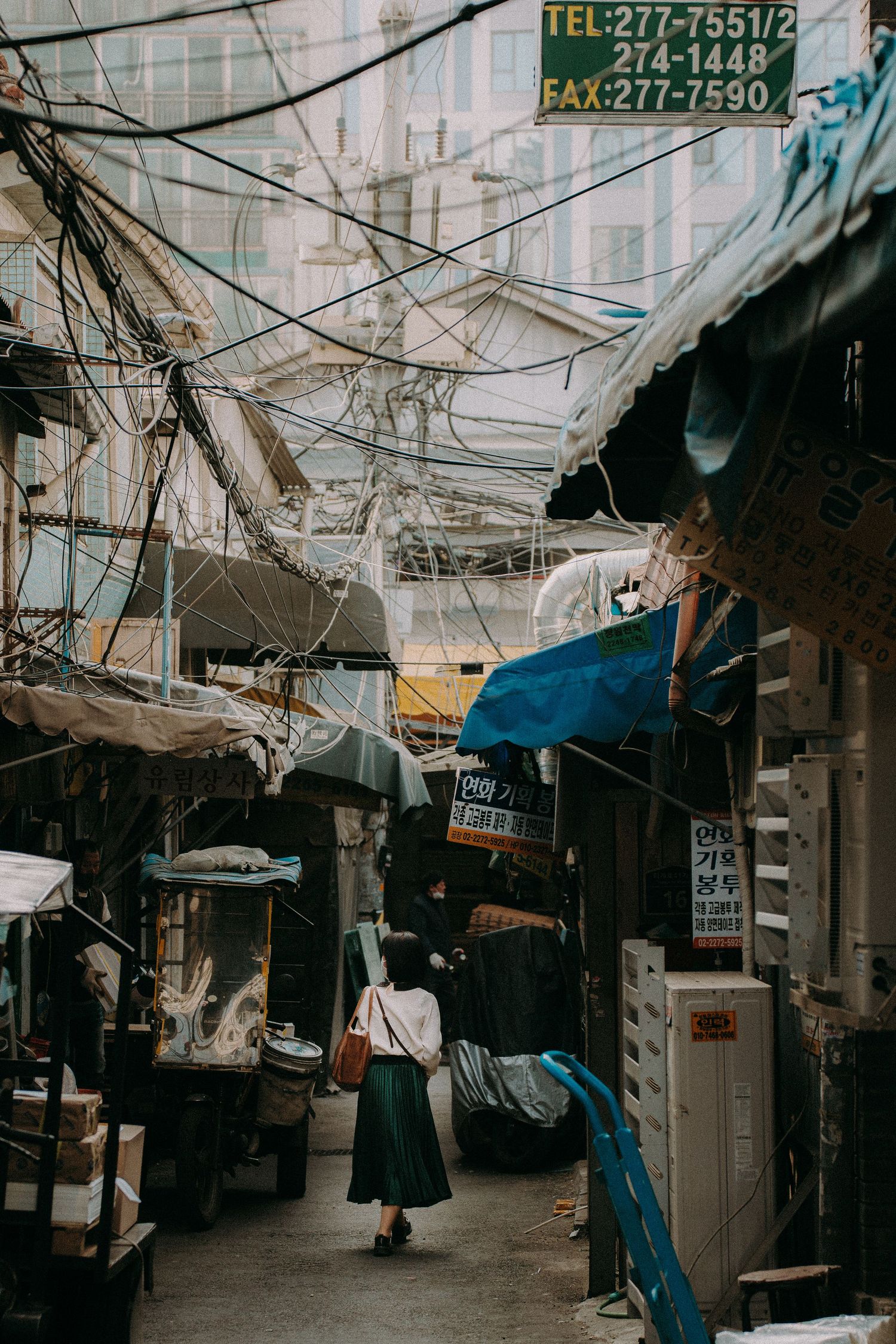 Narrow alley in an urban market with tangled electrical wires and hanging awnings overhead.