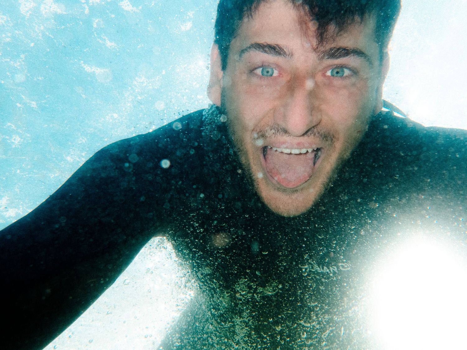 Close-up underwater selfie of someone in a wetsuit with bubbles in turquoise water.