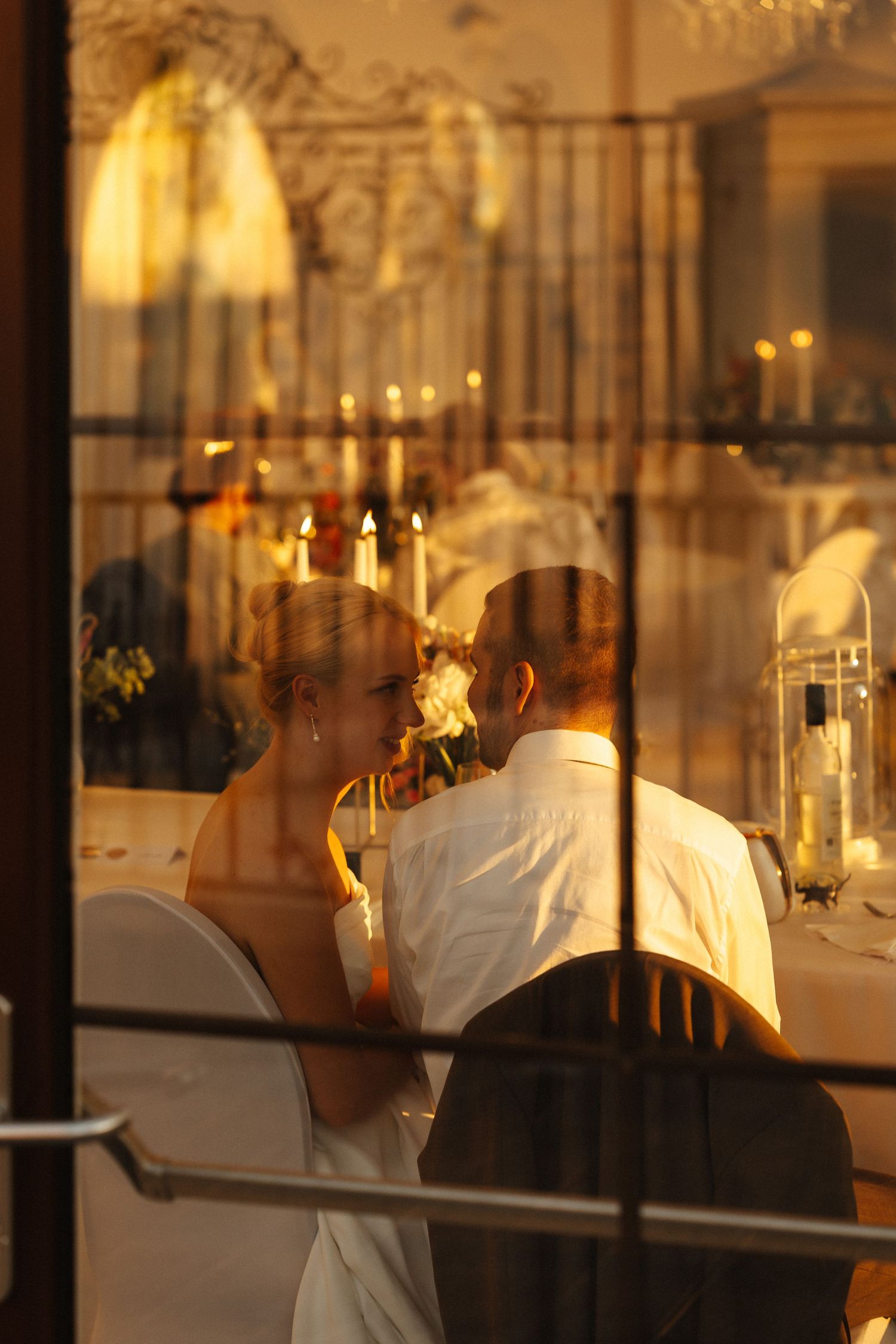 Romantic couple sharing a tender moment through a candlelit window at their wedding reception.