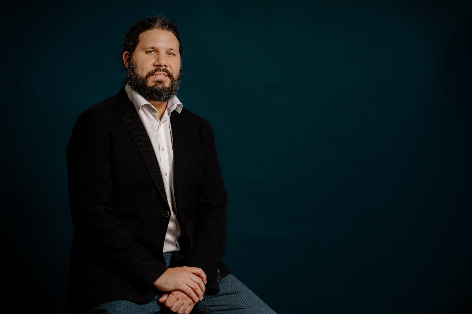 Professional man with beard in black blazer sitting against dark teal background in formal portrait photo.