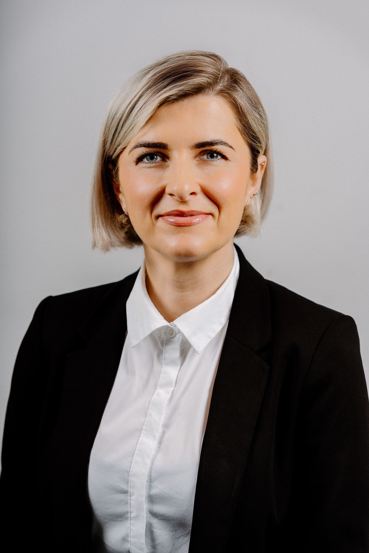 Professional woman in black blazer and white shirt with short blonde hair poses for a corporate headshot.