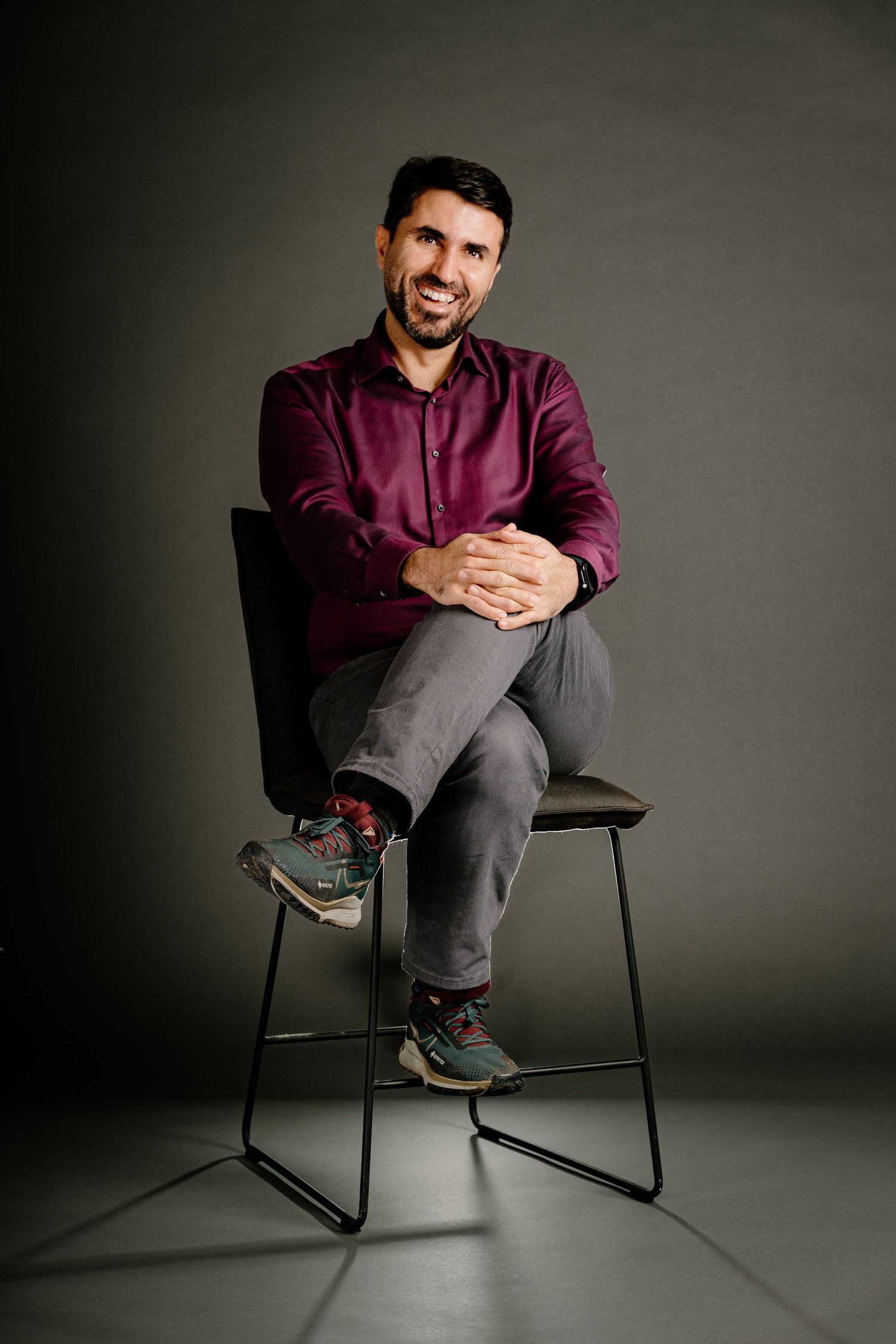 Man in burgundy shirt sitting cross-legged on chair, smiling confidently against gray studio background.