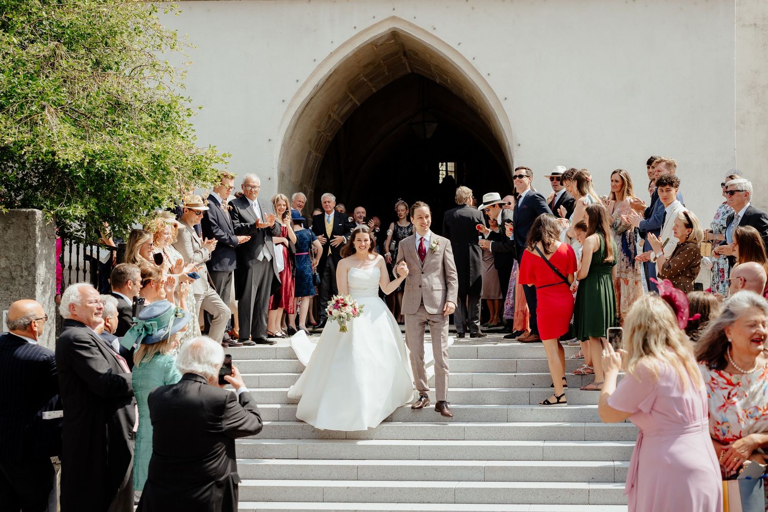 Bride and groom exit church on sunny steps surrounded by smiling wedding guests in elegant attire.