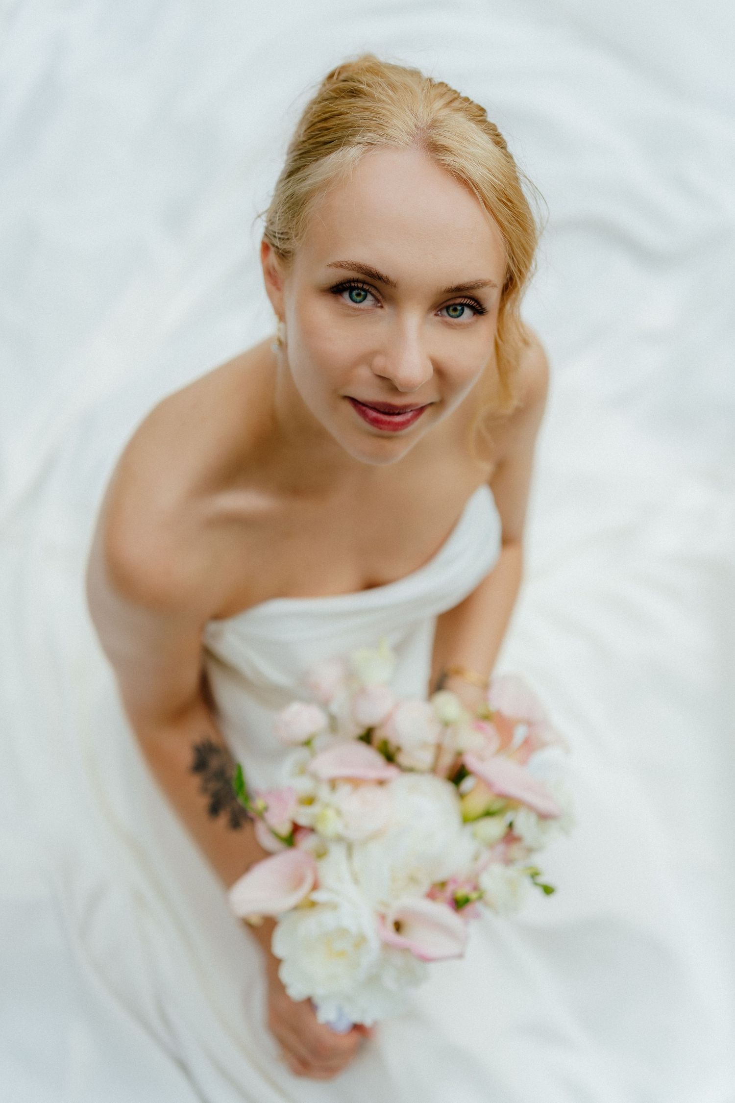 Blonde bride in strapless white gown holding pink and white floral bouquet, looking up at camera with a gentle smile.