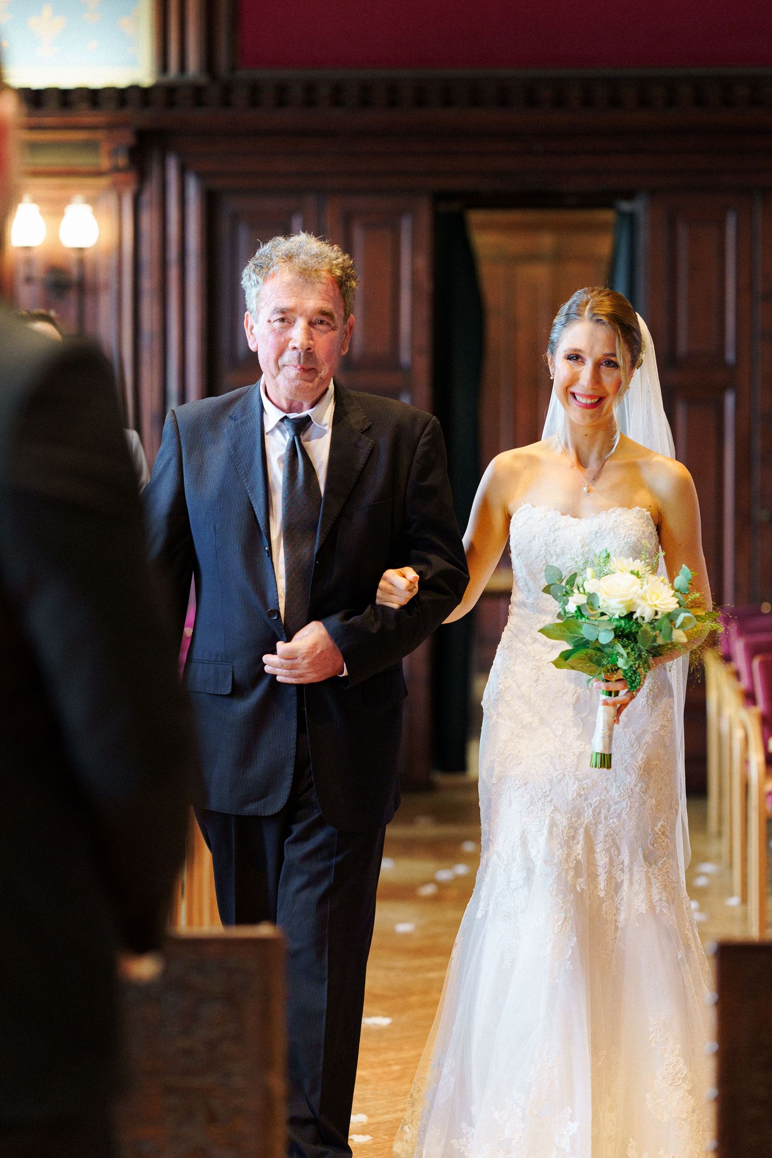 A wedding processional down an elegant wood-paneled hallway with a white bouquet.