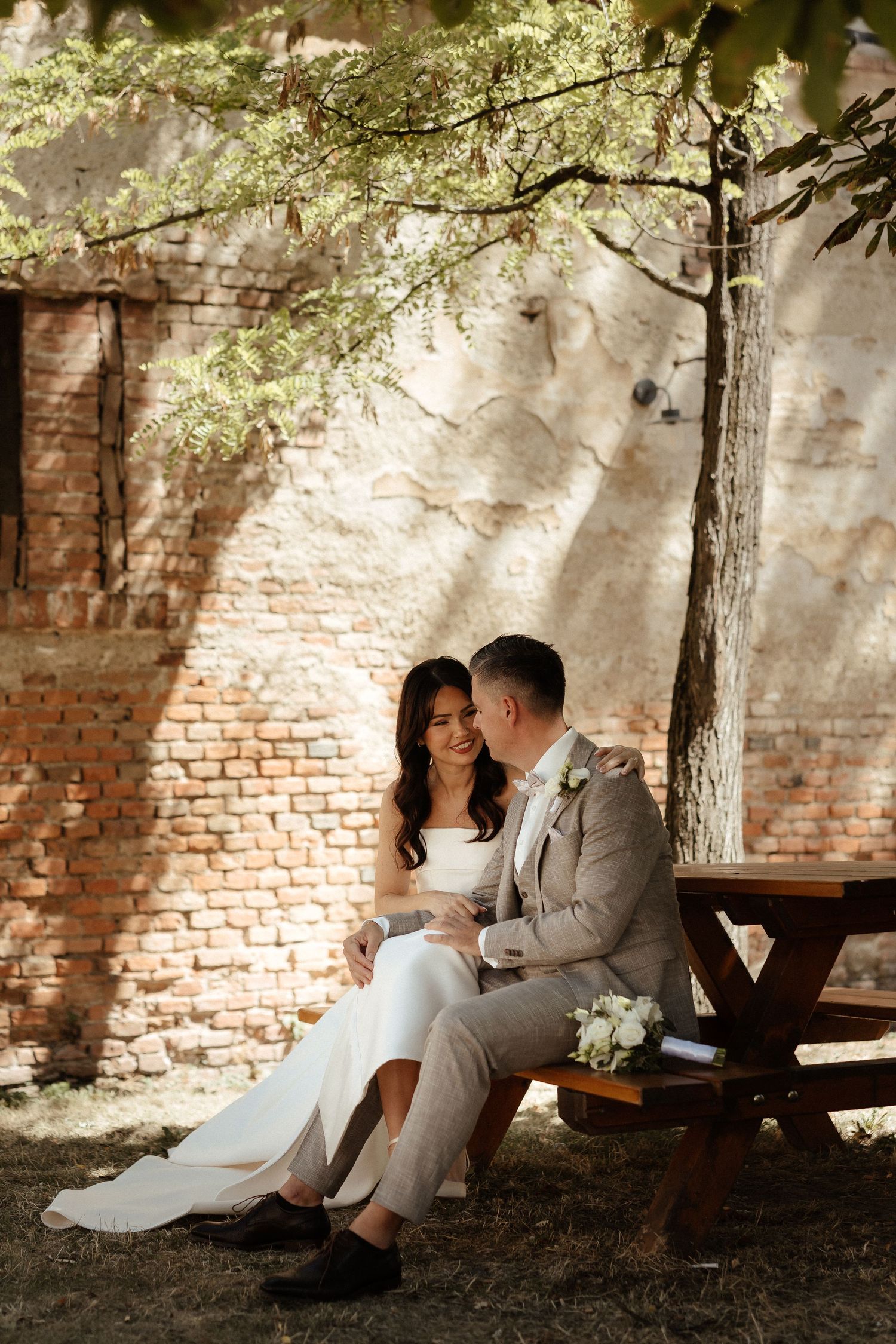 Bride and groom sit together on a bench under a tree against a rustic brick wall in an outdoor wedding photo.