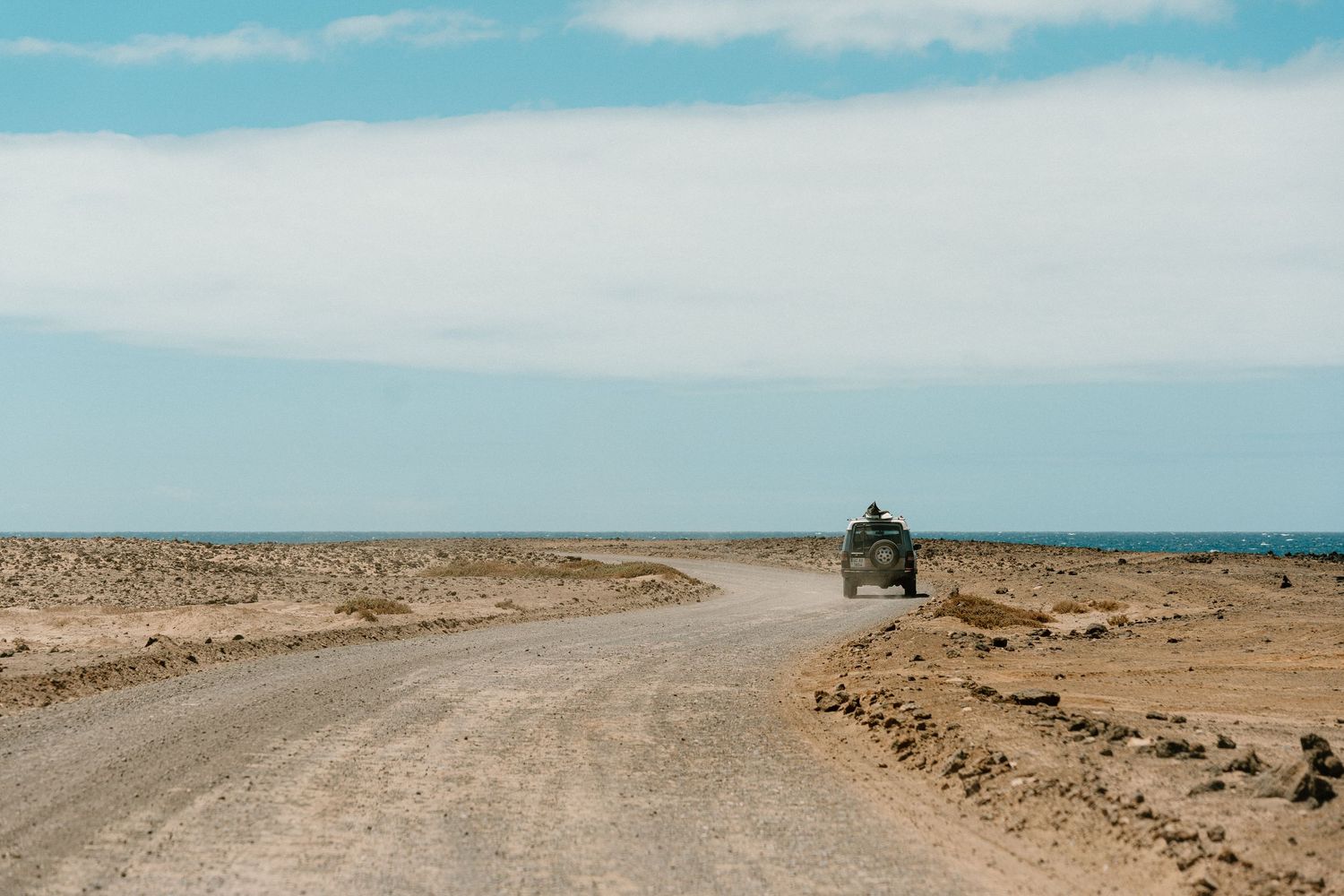 A long empty dirt road stretches across a desert landscape under blue skies.